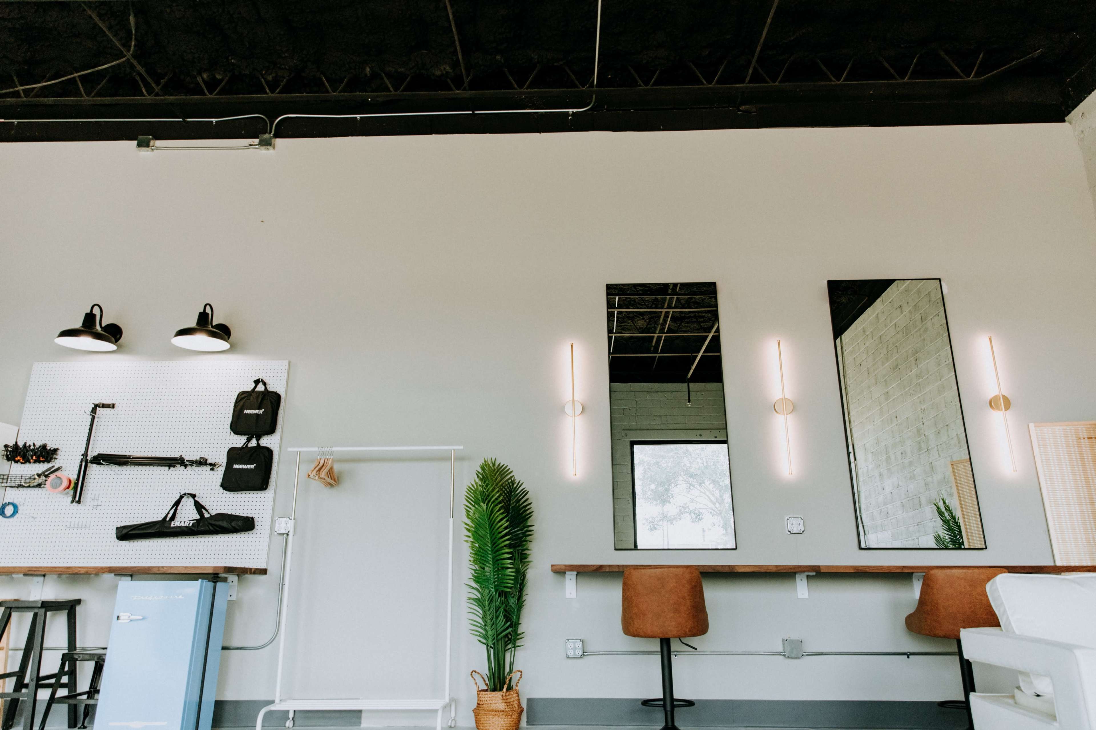 The image shows a modern workspace with long mirrors, pendant lights, a green plant, and two barstools positioned at a wooden counter against a neutral wall.