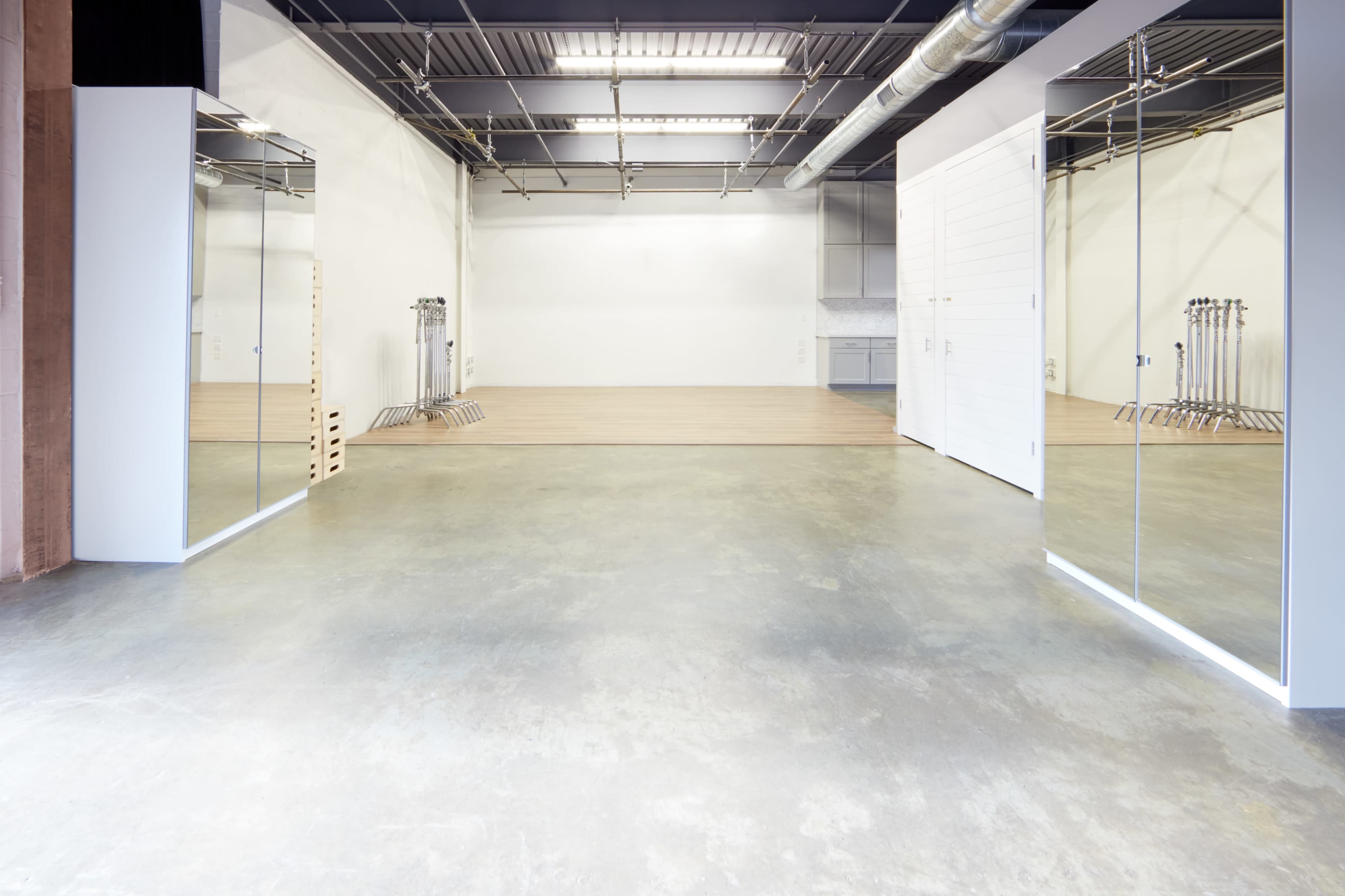 An empty room with concrete flooring, large mirrors on the walls, and wooden flooring in the background.