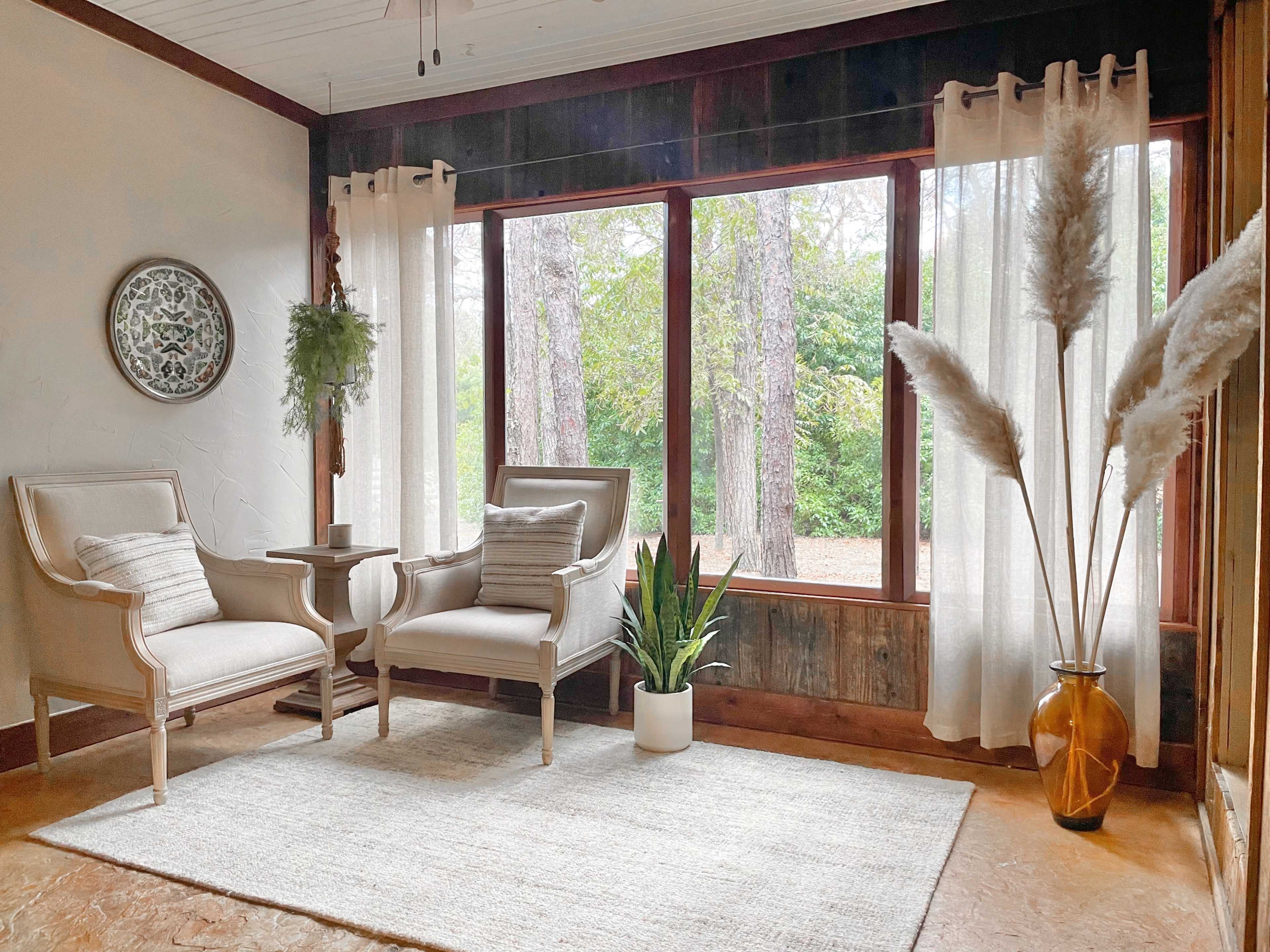 A cozy sitting area features two armchairs, a small table, a plant, and large windows with sheer curtains, allowing natural light to illuminate the space.