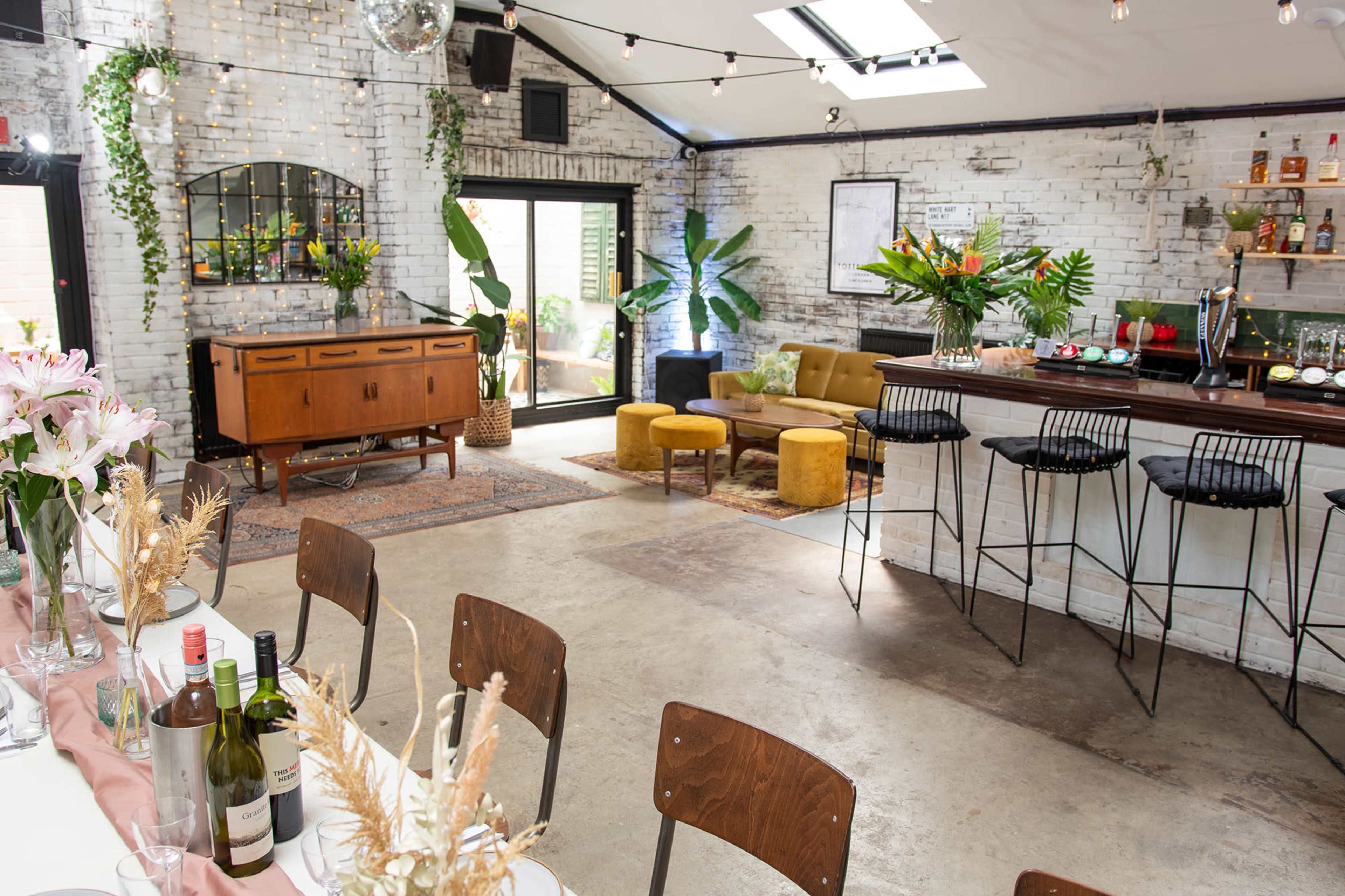 A modern, well-lit event space featuring a bar, seating areas, and decorative plants against a backdrop of white brick walls.