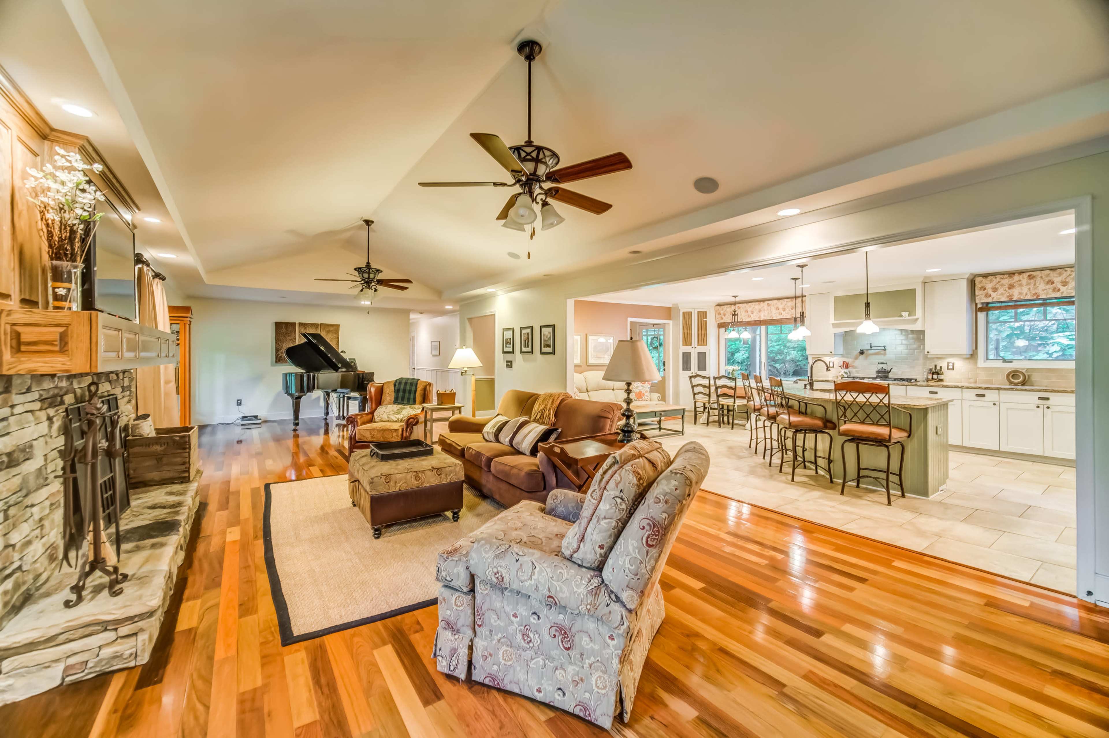 The image shows a spacious living room with a stone fireplace, hardwood floors, and a view of an open kitchen in the background.