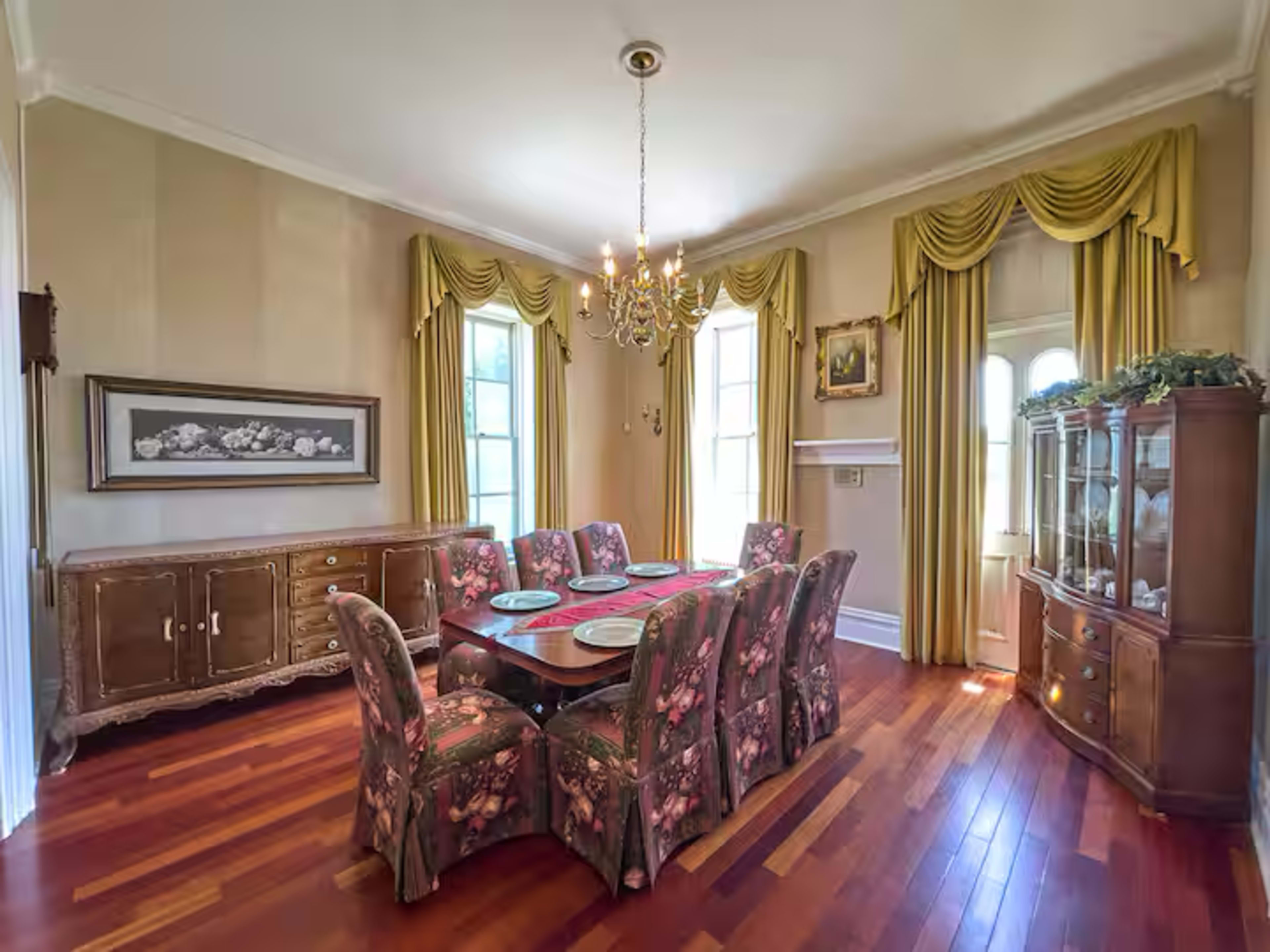 The image shows a traditional dining room with a wooden table surrounded by chairs upholstered in floral fabric, draped curtains, and a chandelier overhead.