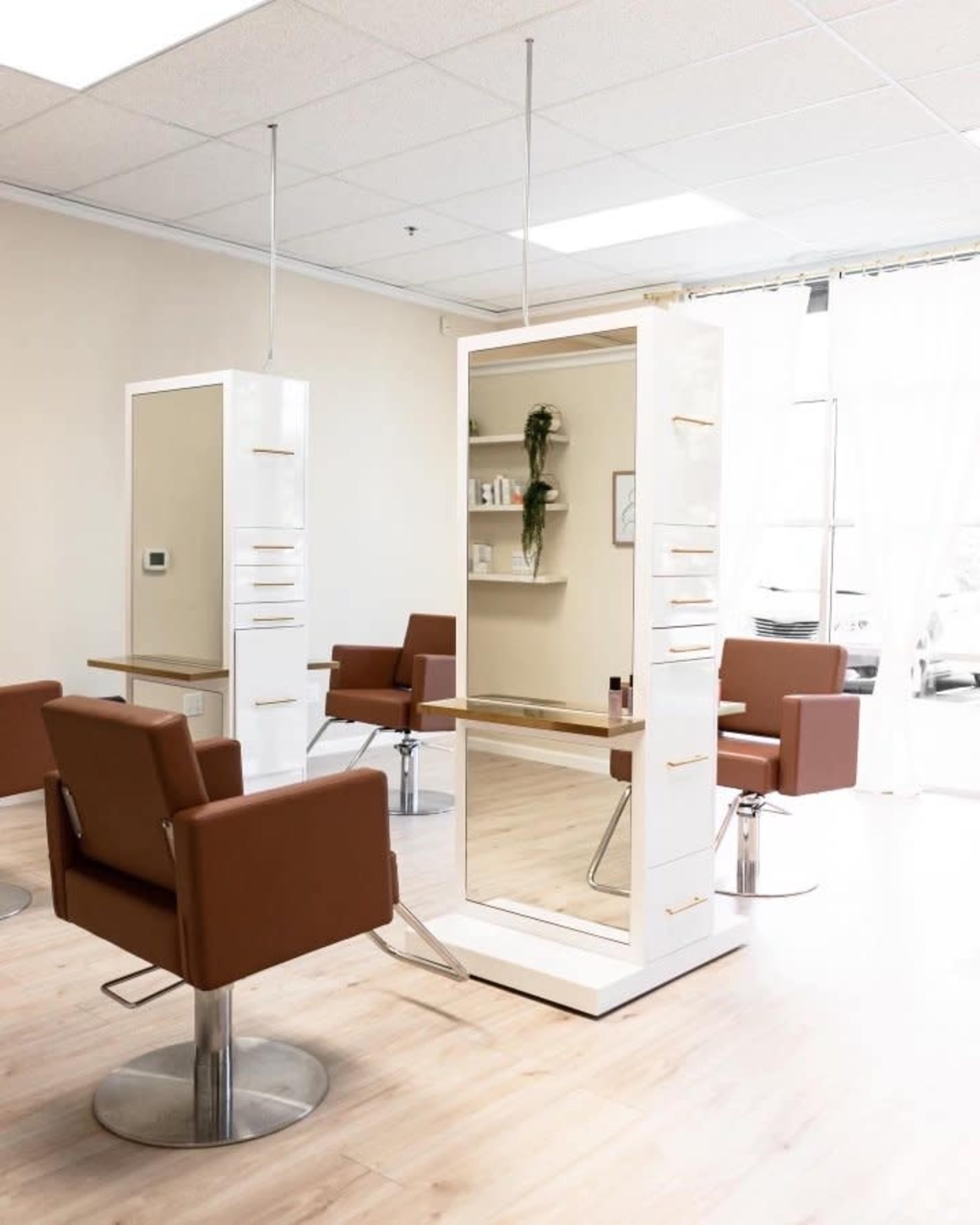 A modern hair salon with two styling stations featuring large mirrors and brown chairs arranged around a light-colored wooden floor.