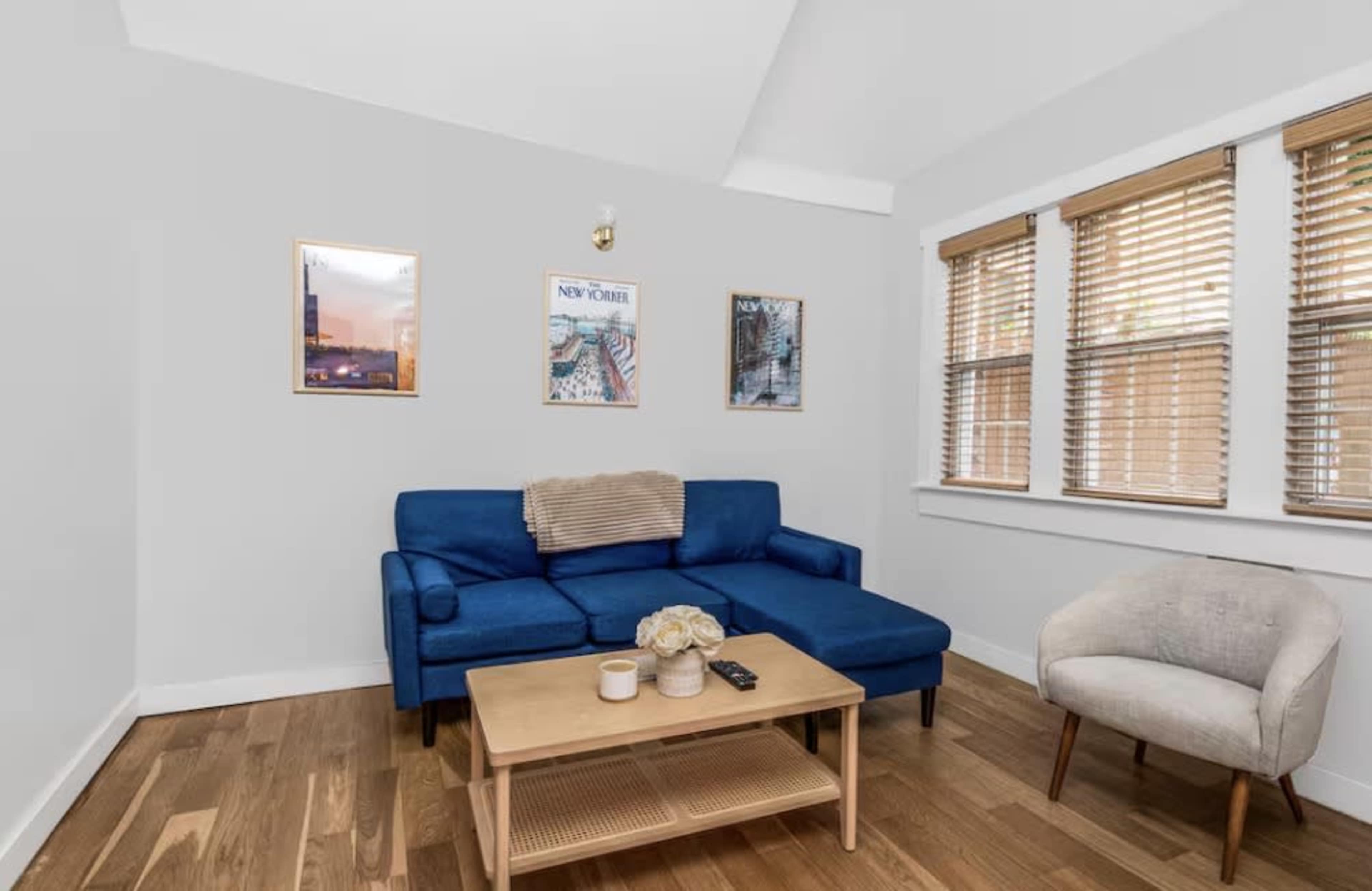 The image shows a living room featuring a blue sofa, a light-colored armchair, a wooden coffee table, and framed artwork on the walls.