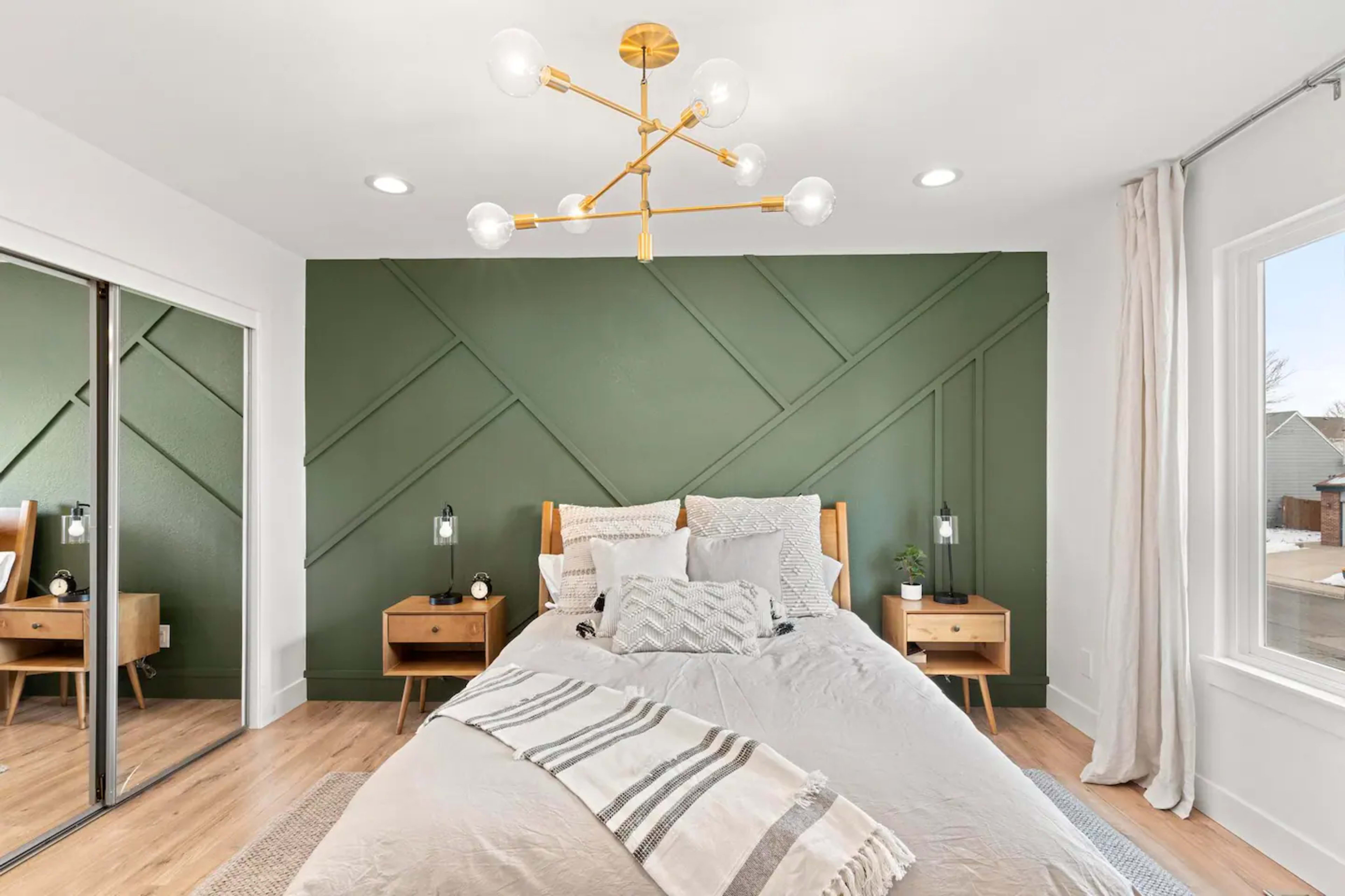 A well-designed bedroom features a green accent wall, a bed with decorative pillows, two bedside tables with lamps, and a modern light fixture.