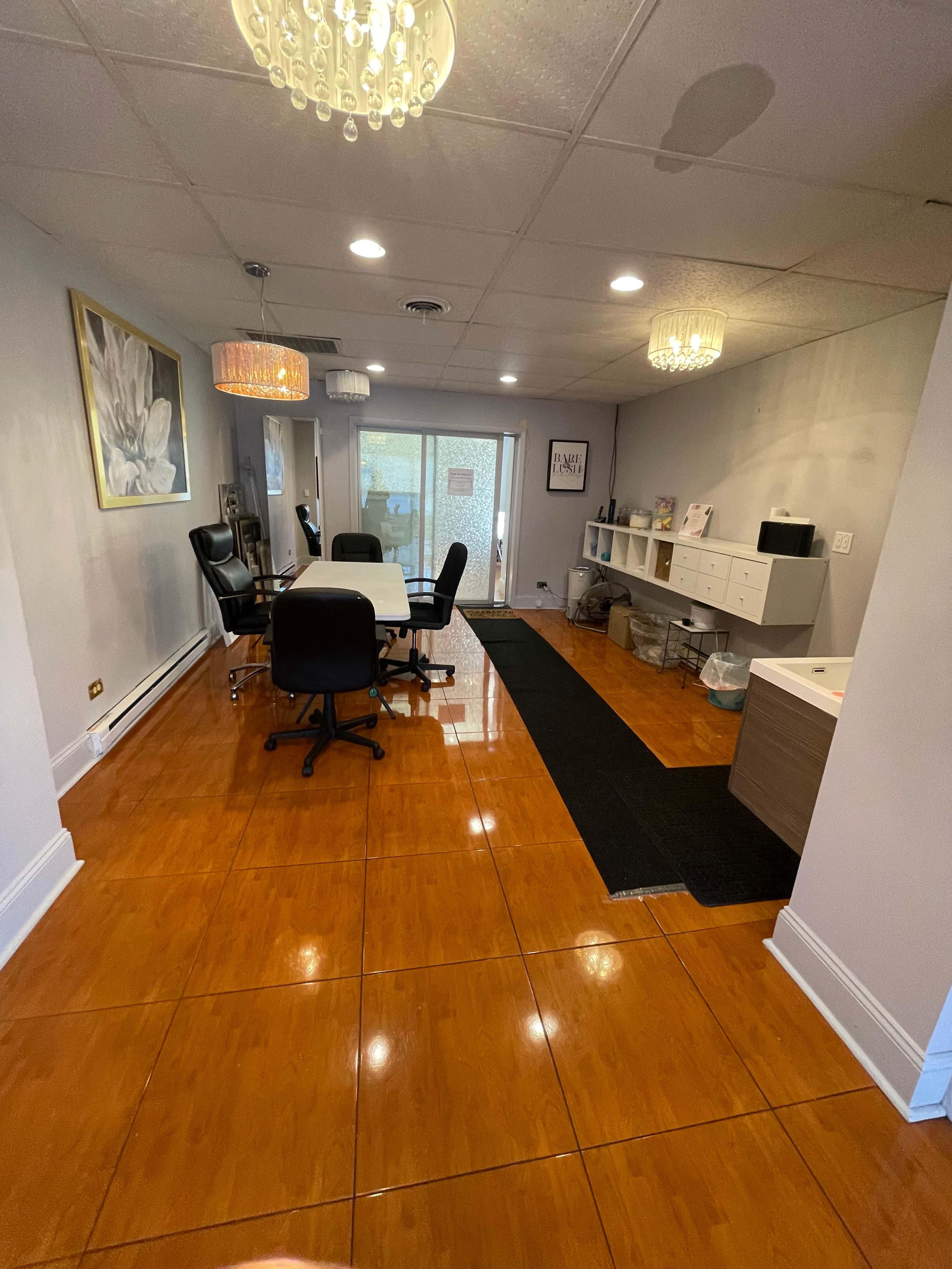 A spacious office area with a long table, multiple chairs, a side cabinet, and large windows leading to an outdoor space, all featuring polished tile flooring.