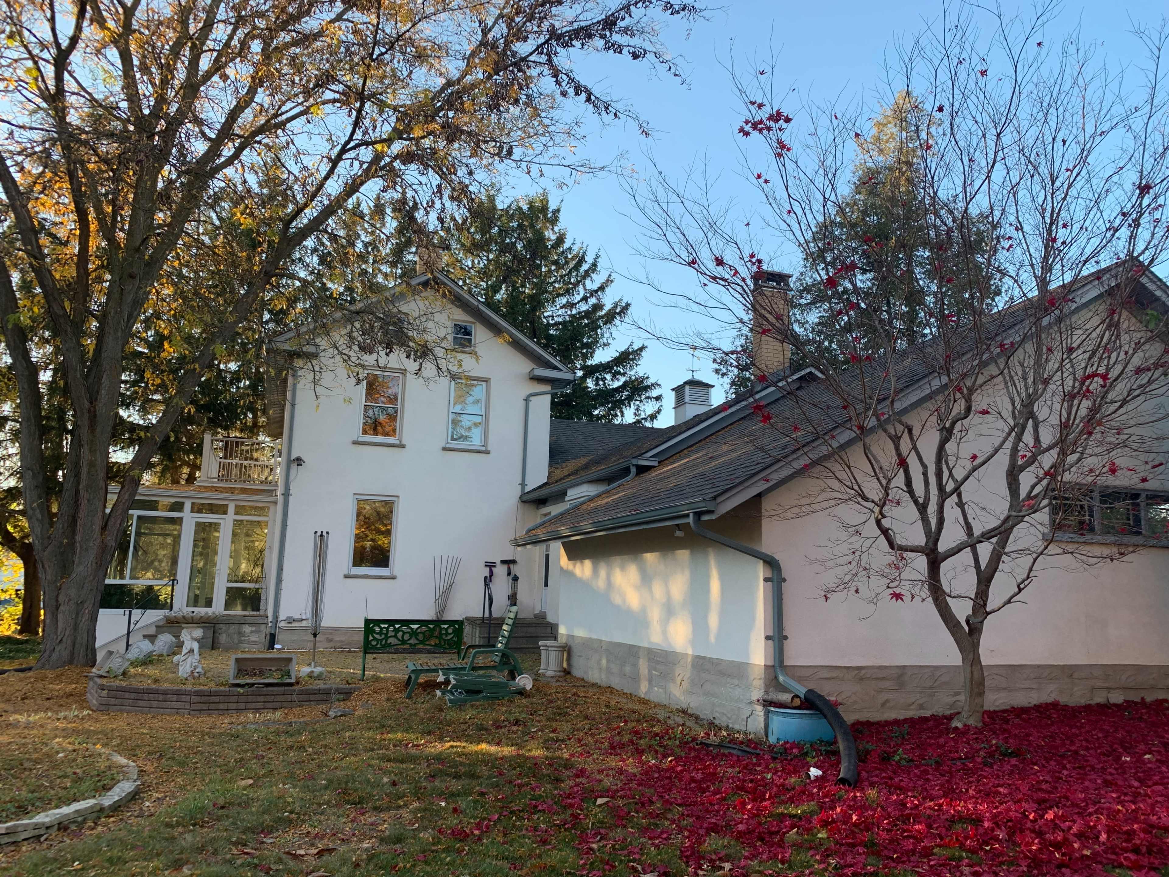 A two-story house with a green balcony and a side garden is surrounded by trees and decorative landscaping, featuring fallen red leaves.