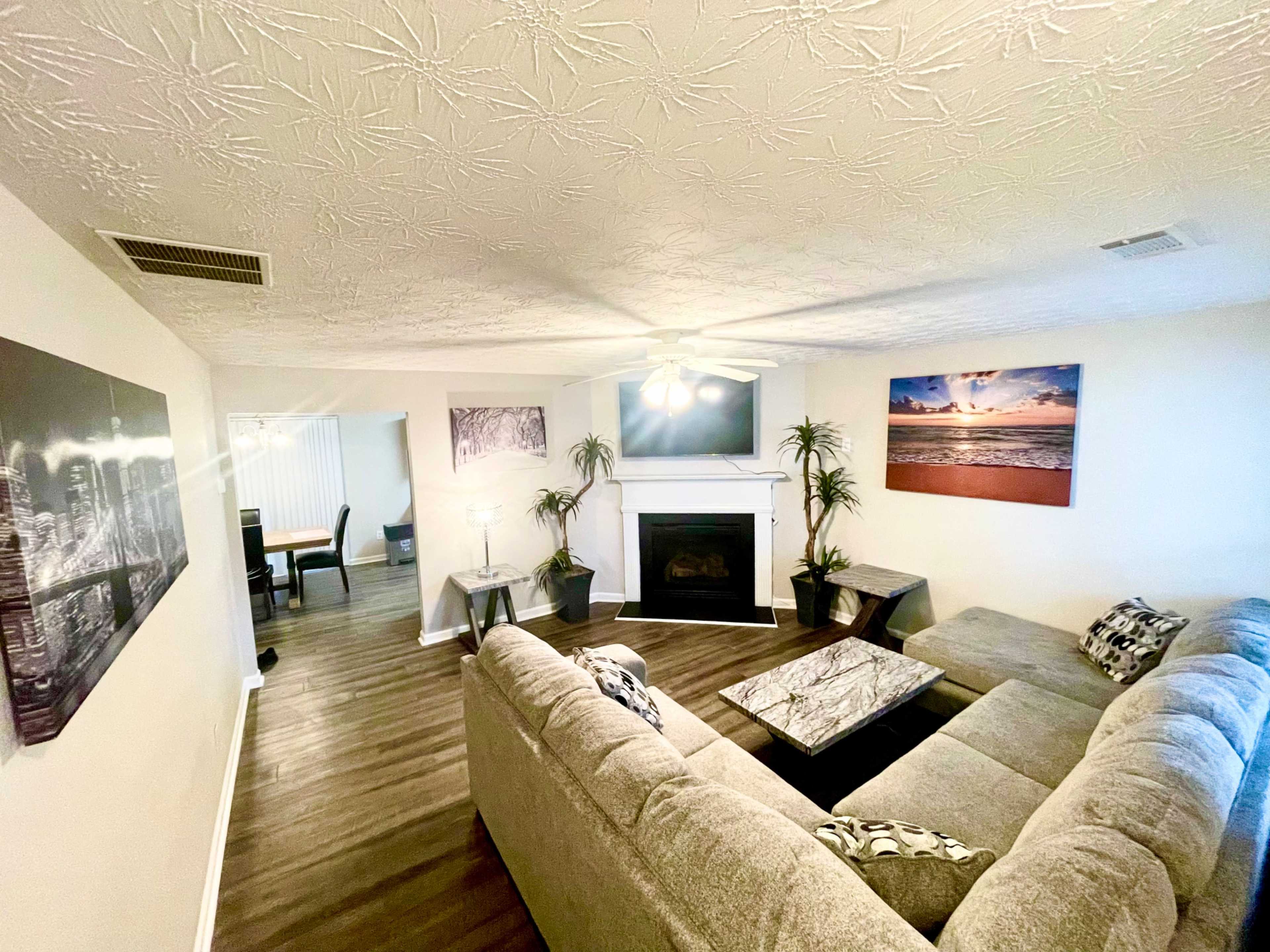 The living room features a sectional sofa, a coffee table, and a fireplace, with framed artwork and indoor plants decorating the space.