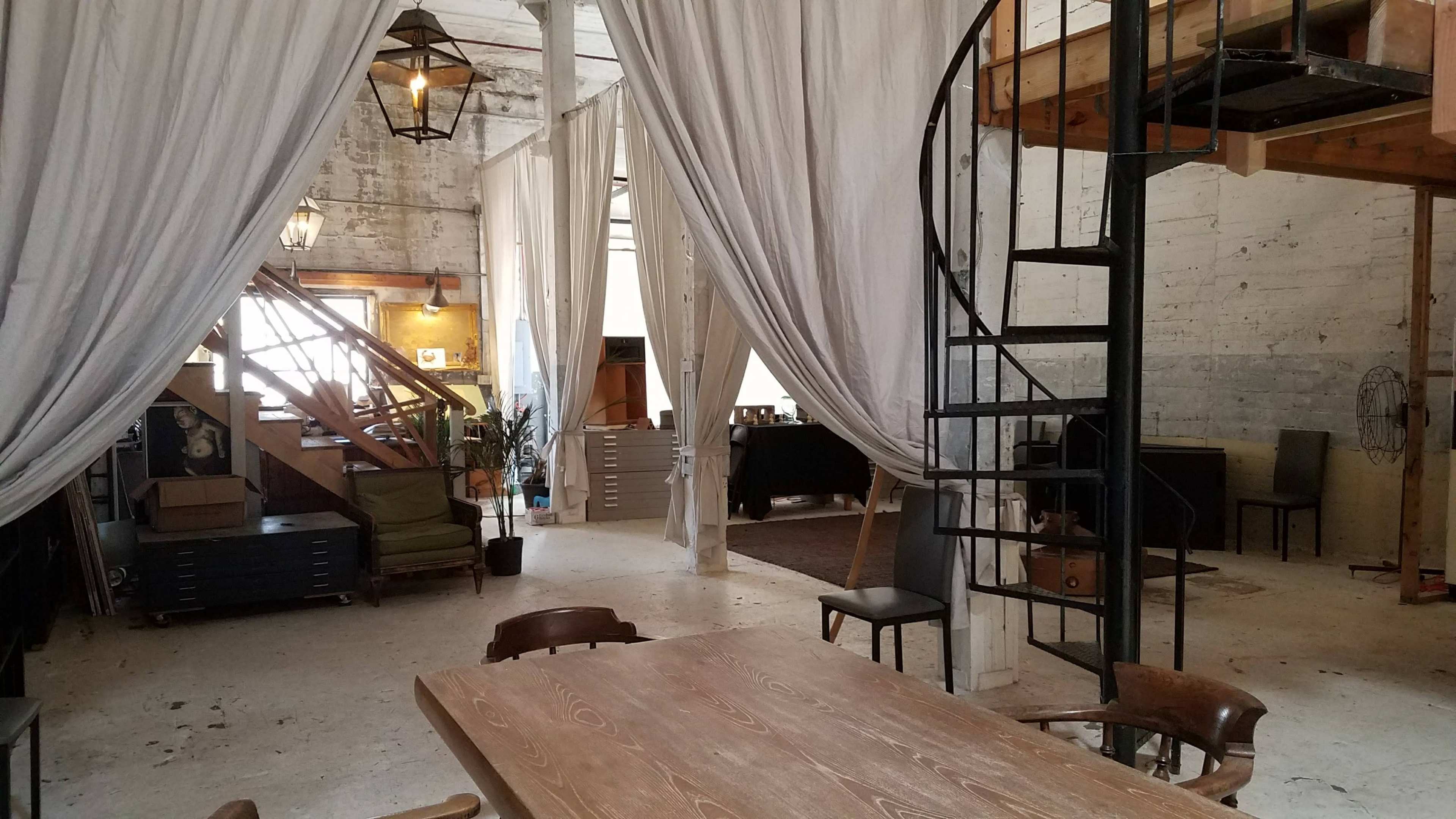 A spacious, industrial-style room featuring a wooden table in the foreground, a spiral staircase to the right, and separated areas by white curtains.