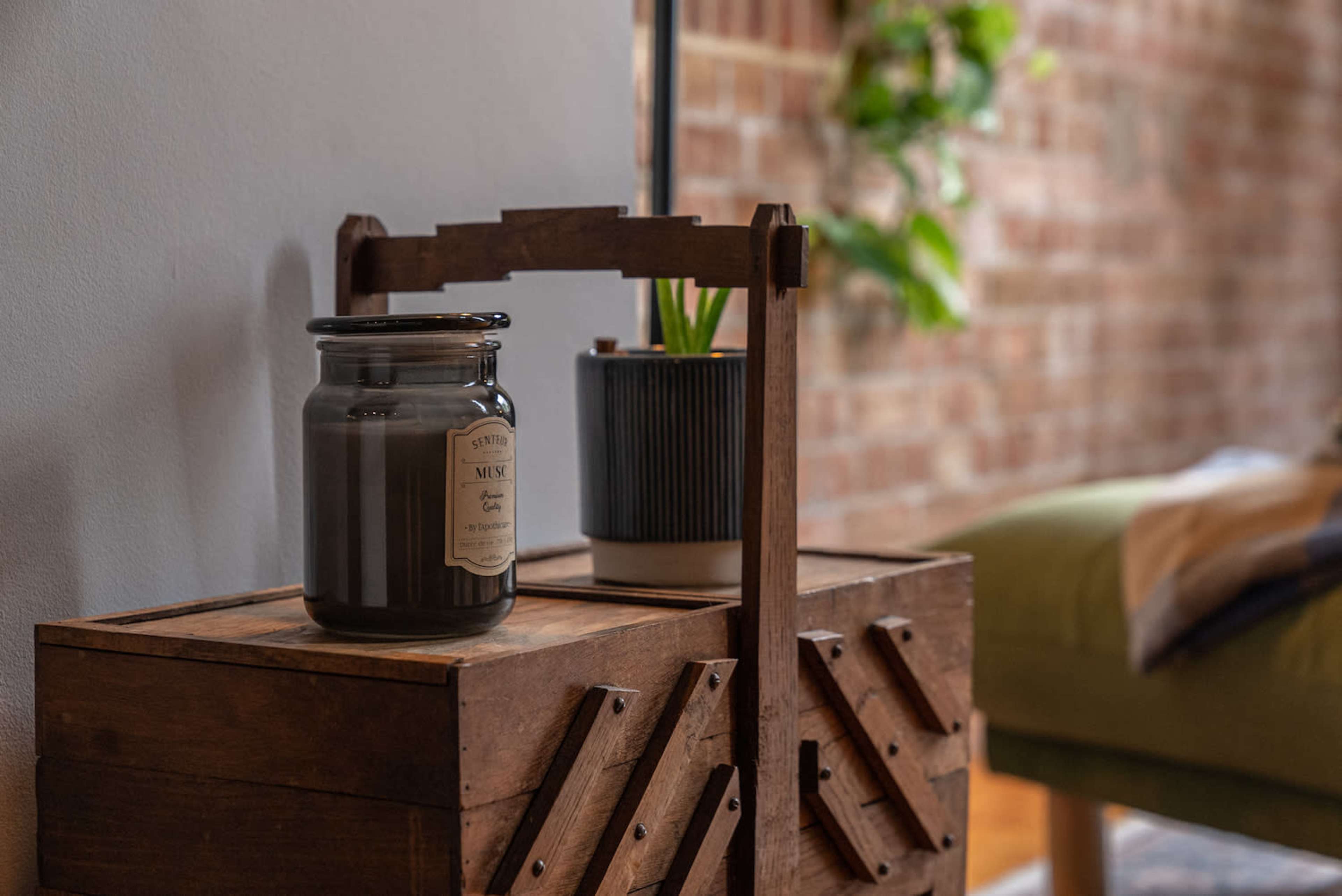 A wooden side table holds a candle jar and a small potted plant in a brick-walled room.