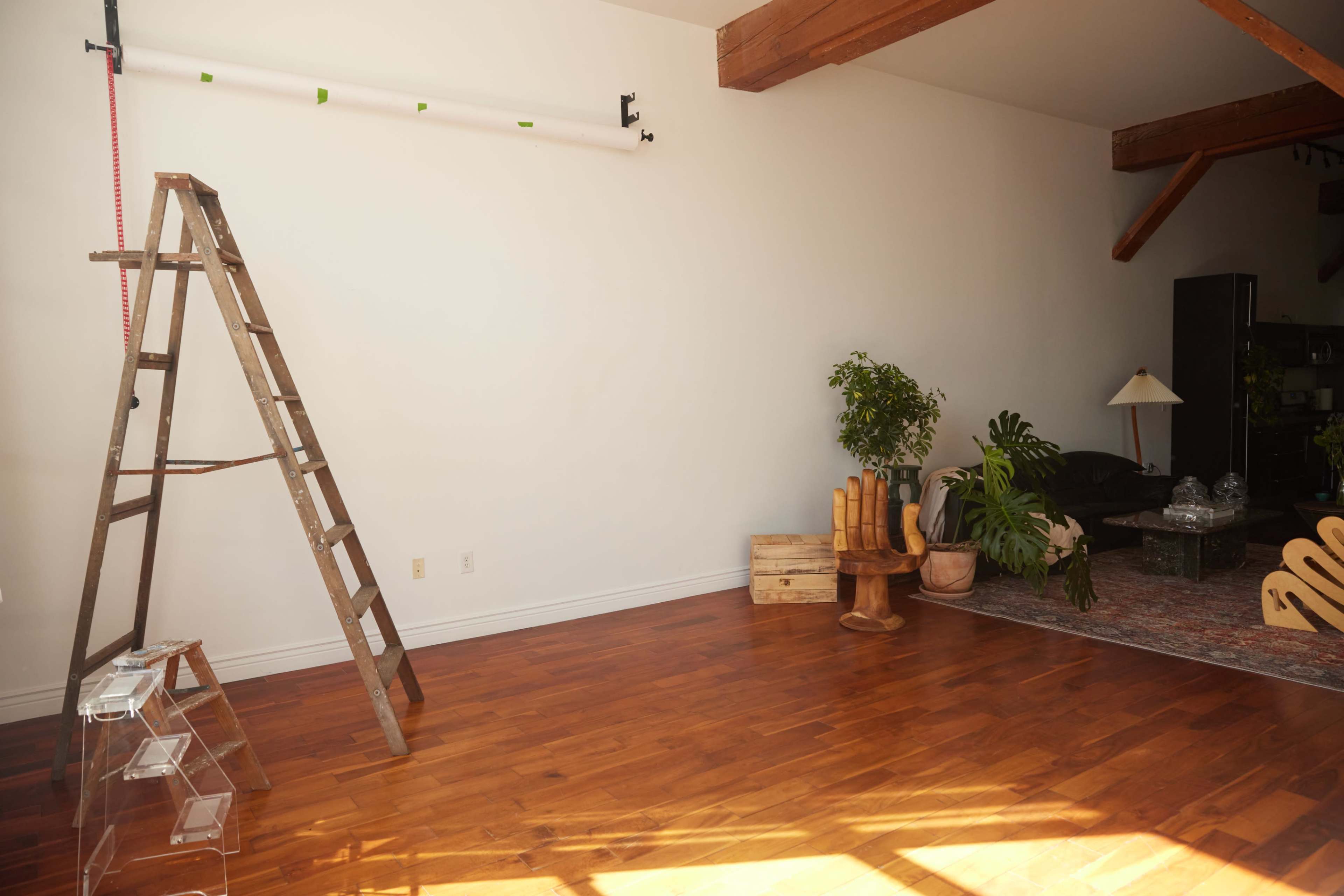 The image shows a spacious room with wooden flooring, a ladder against a wall, plants in decorative pots, and a seating area with a rug.