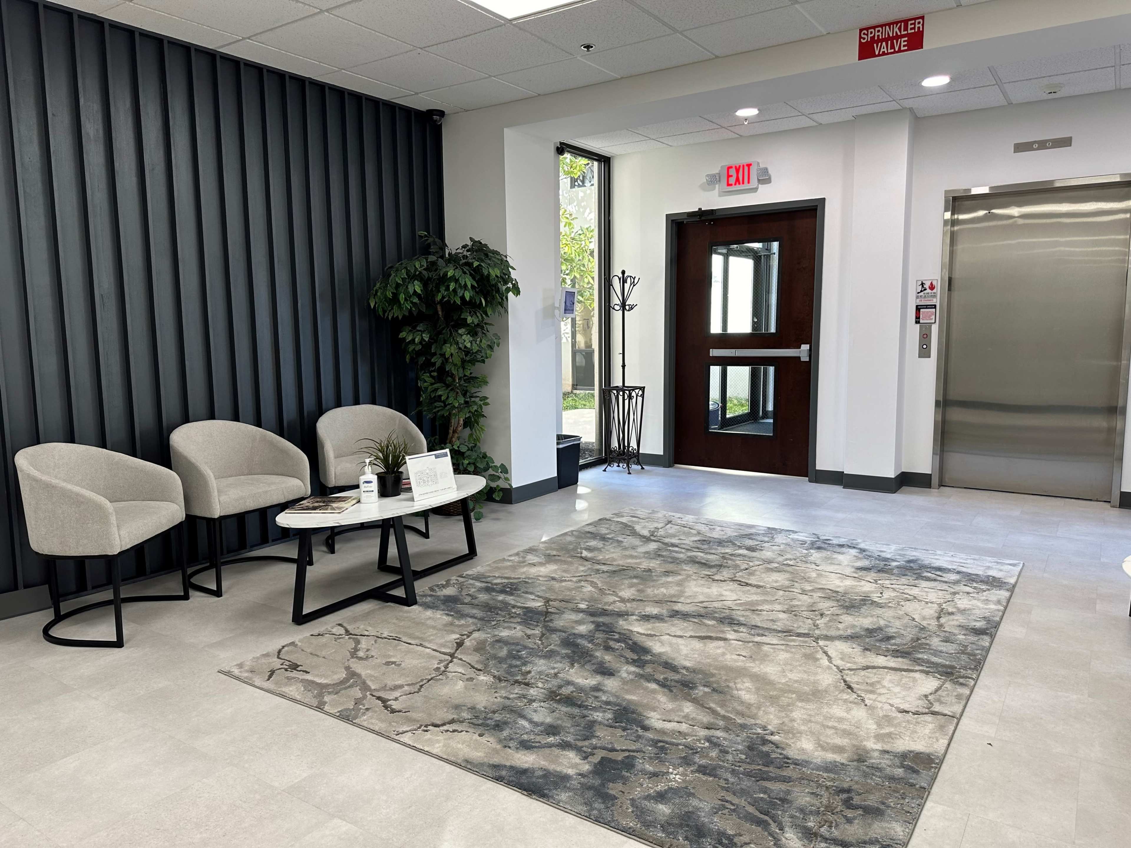 The image shows a modern lobby area with two upholstered chairs, a small table, a potted plant, and an elevator in the background.