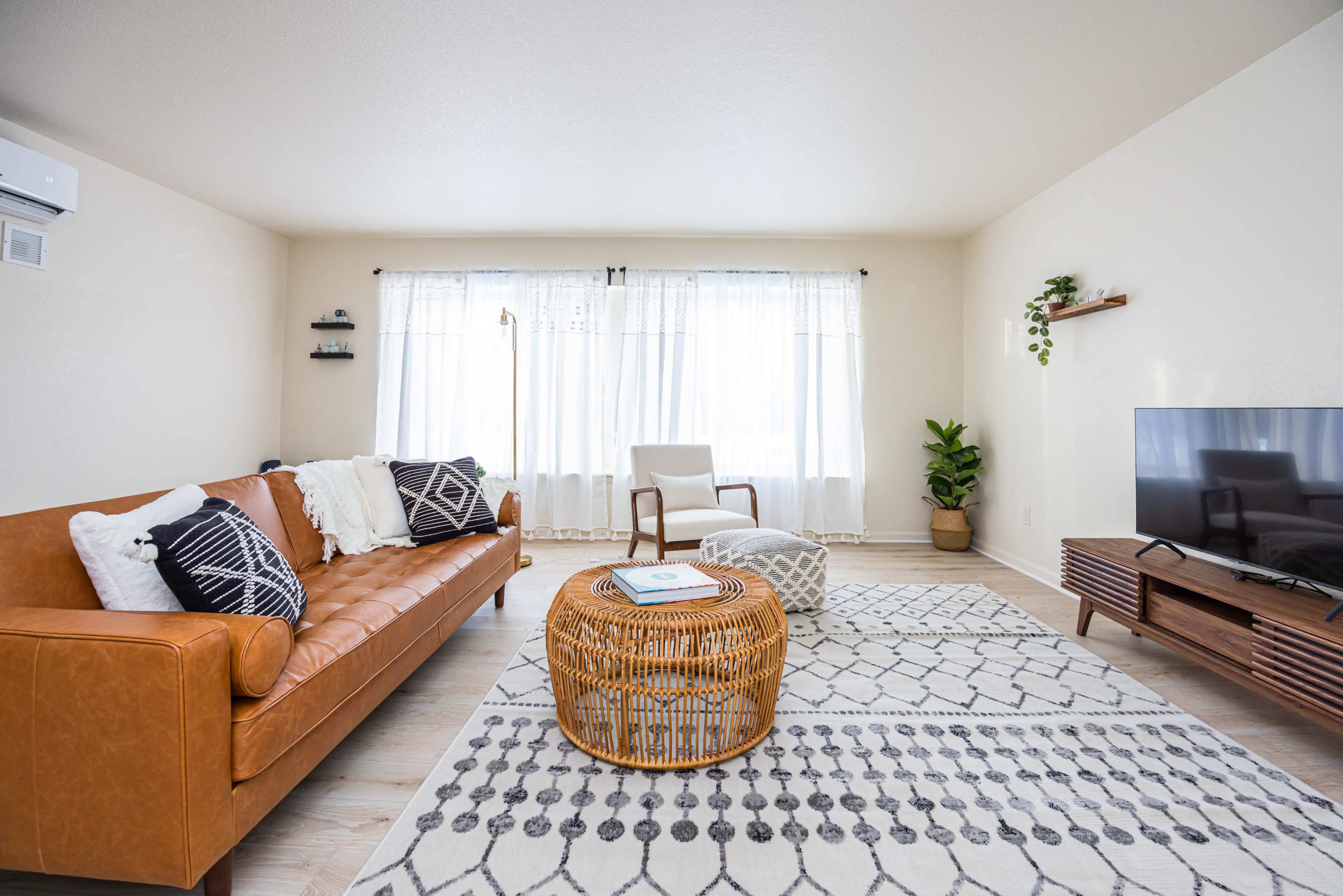 A bright and airy living room features a brown leather sofa, a round woven coffee table, and large windows with sheer curtains.