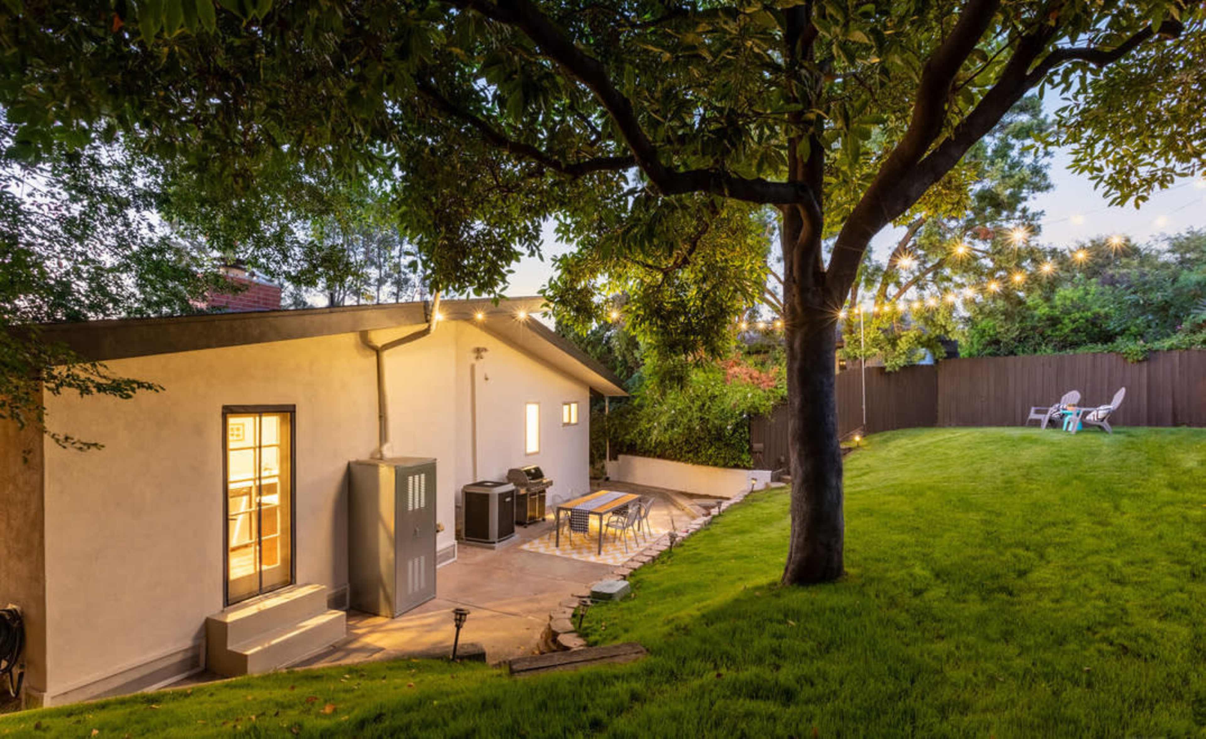 A well-lit backyard features a patio with outdoor furniture, surrounded by lush grass and trees, and string lights hanging above.