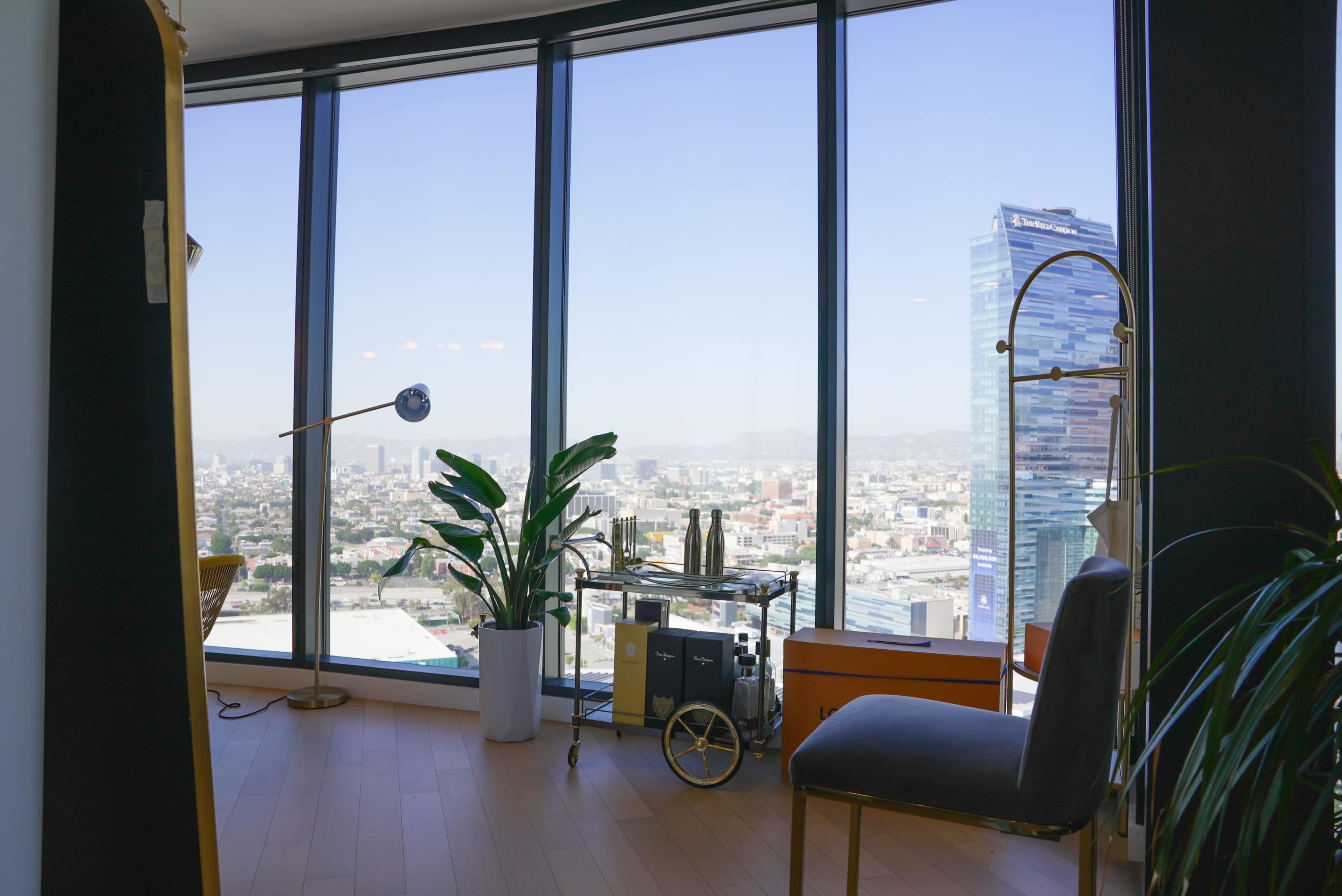 The image shows a modern office space featuring large windows with a city view, a plant, a bar cart, and a chair.