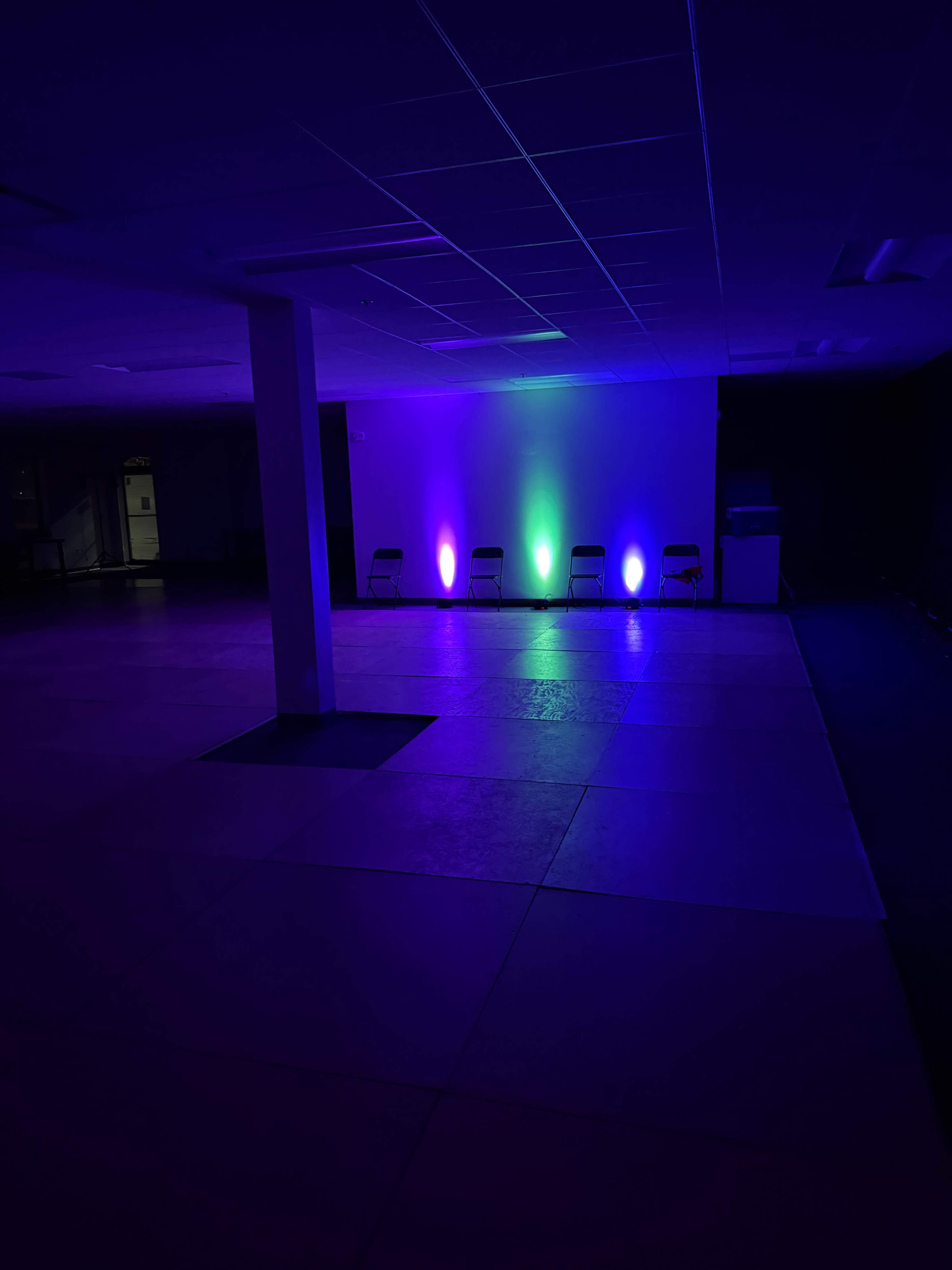 The scene shows a dimly lit interior room with a large empty floor, two rows of chairs along one wall, and colorful lights illuminating the space.