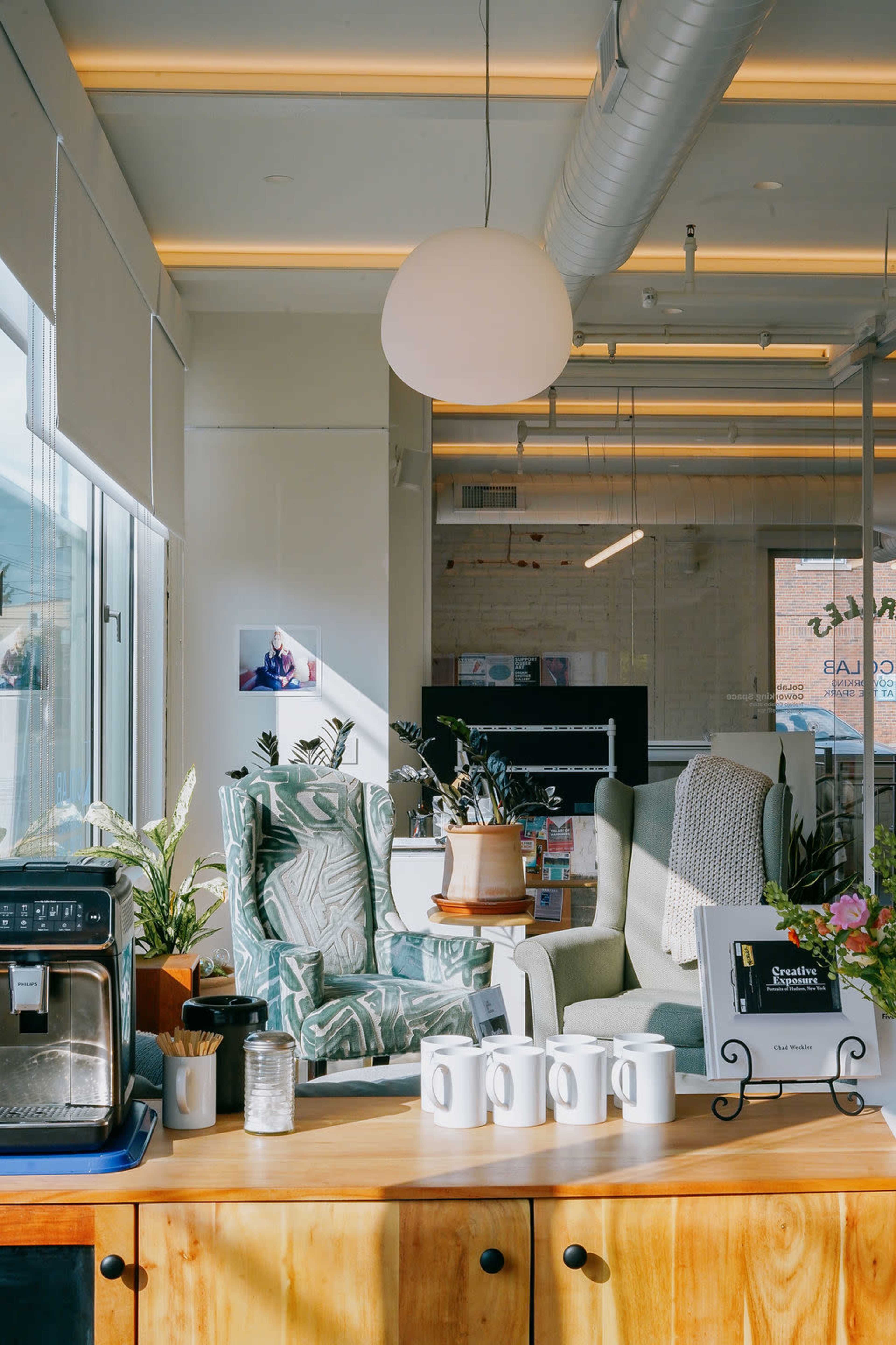The image shows a cozy café interior featuring two patterned armchairs, a coffee machine, and a countertop with several mugs.