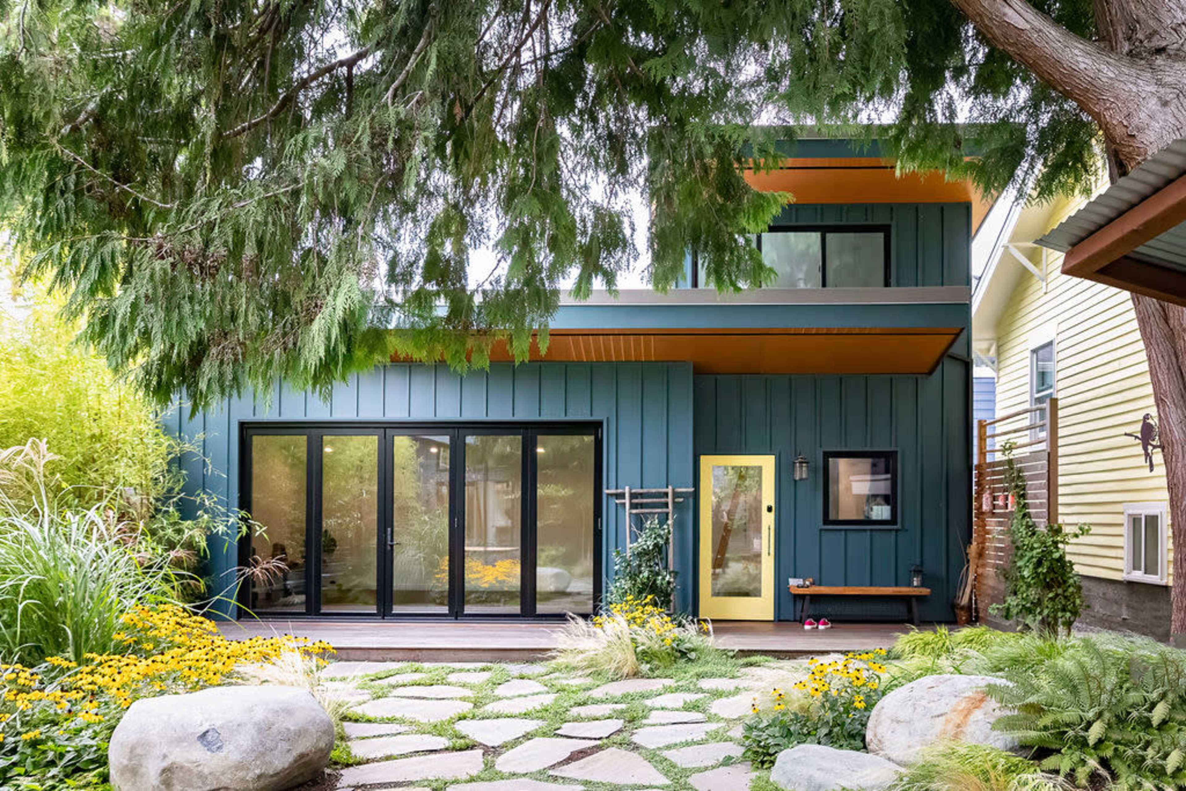 A modern two-story house with dark green siding, large glass doors, and a yellow front door is surrounded by lush greenery and blooming flowers.