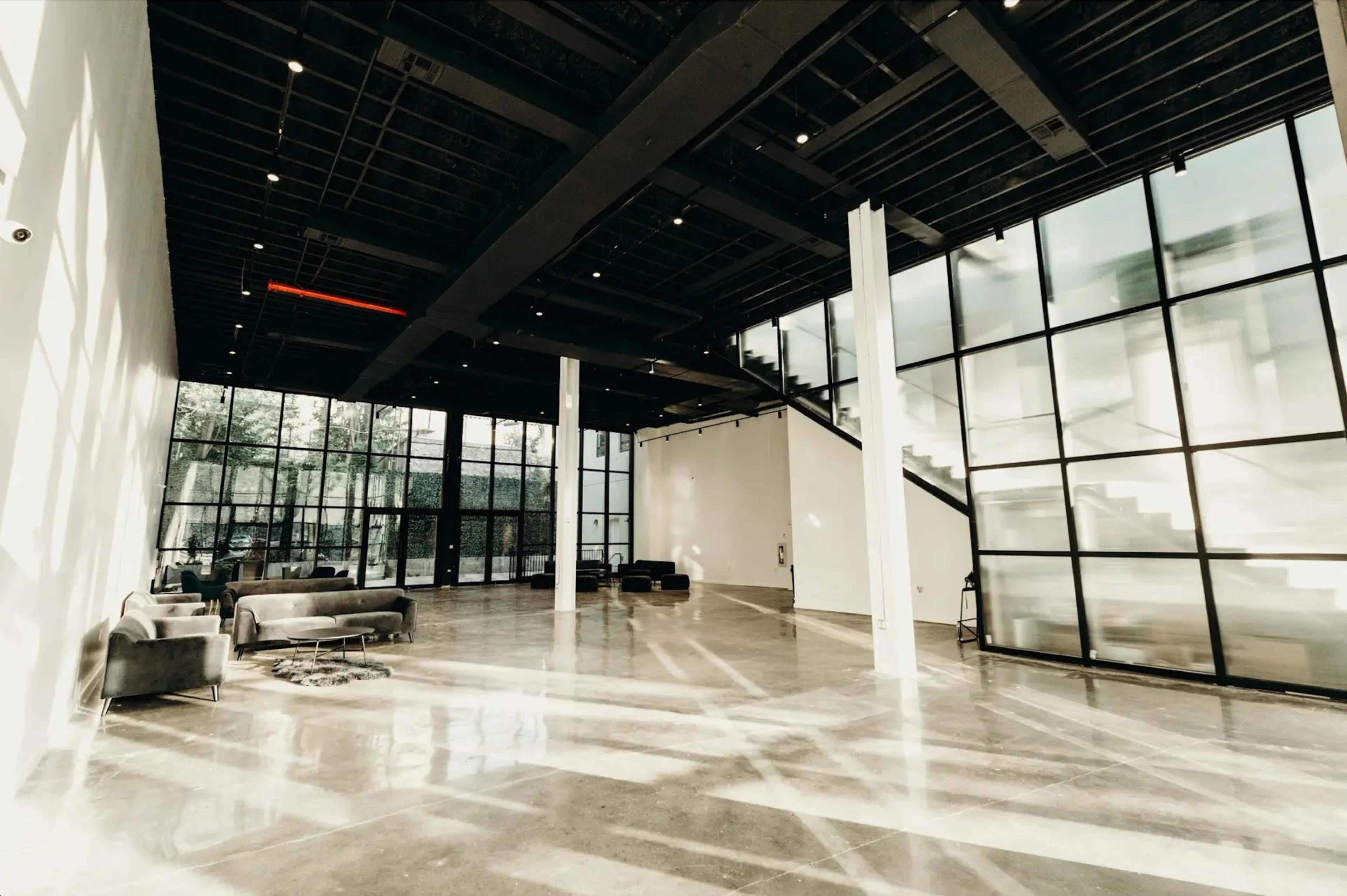 A spacious, modern interior featuring concrete floors, large glass windows, and minimalistic furniture arranged in an open area.