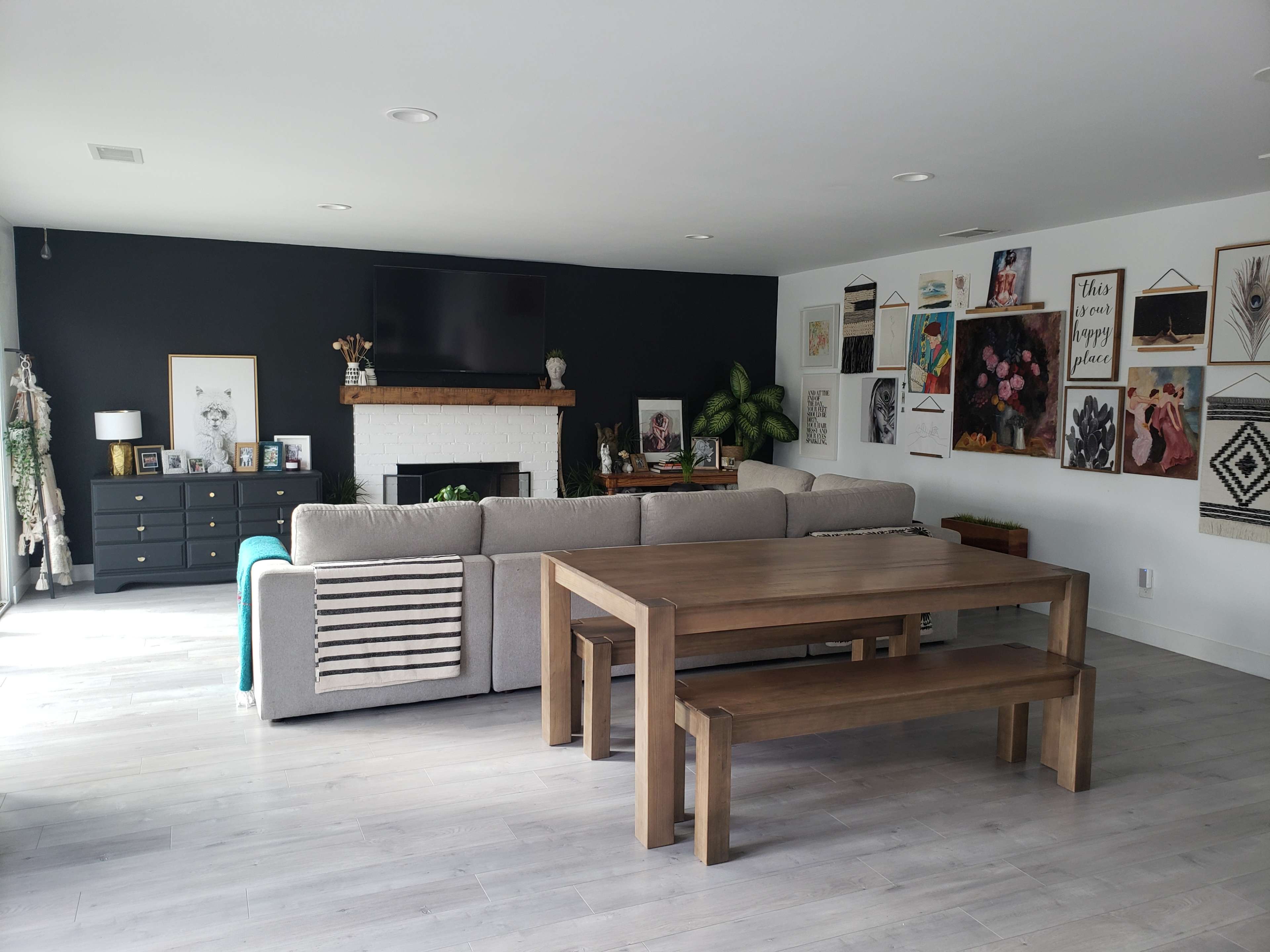 A modern living room features a large gray sectional sofa, a wooden dining table with benches, and a wall adorned with various framed artworks.