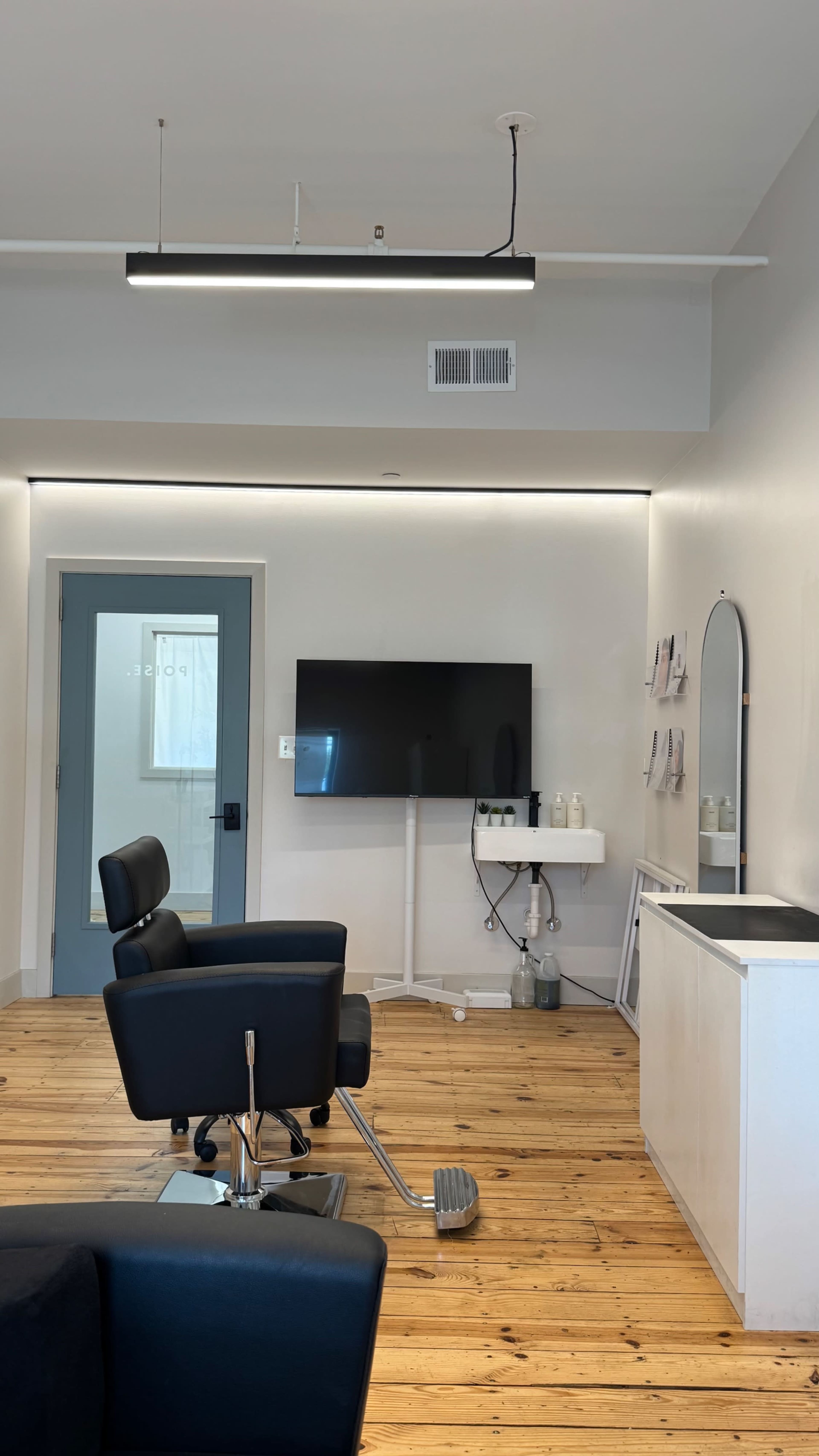 The image shows a modern hair salon interior featuring a chair, a television mounted on the wall, a styling station, and a door leading outside.