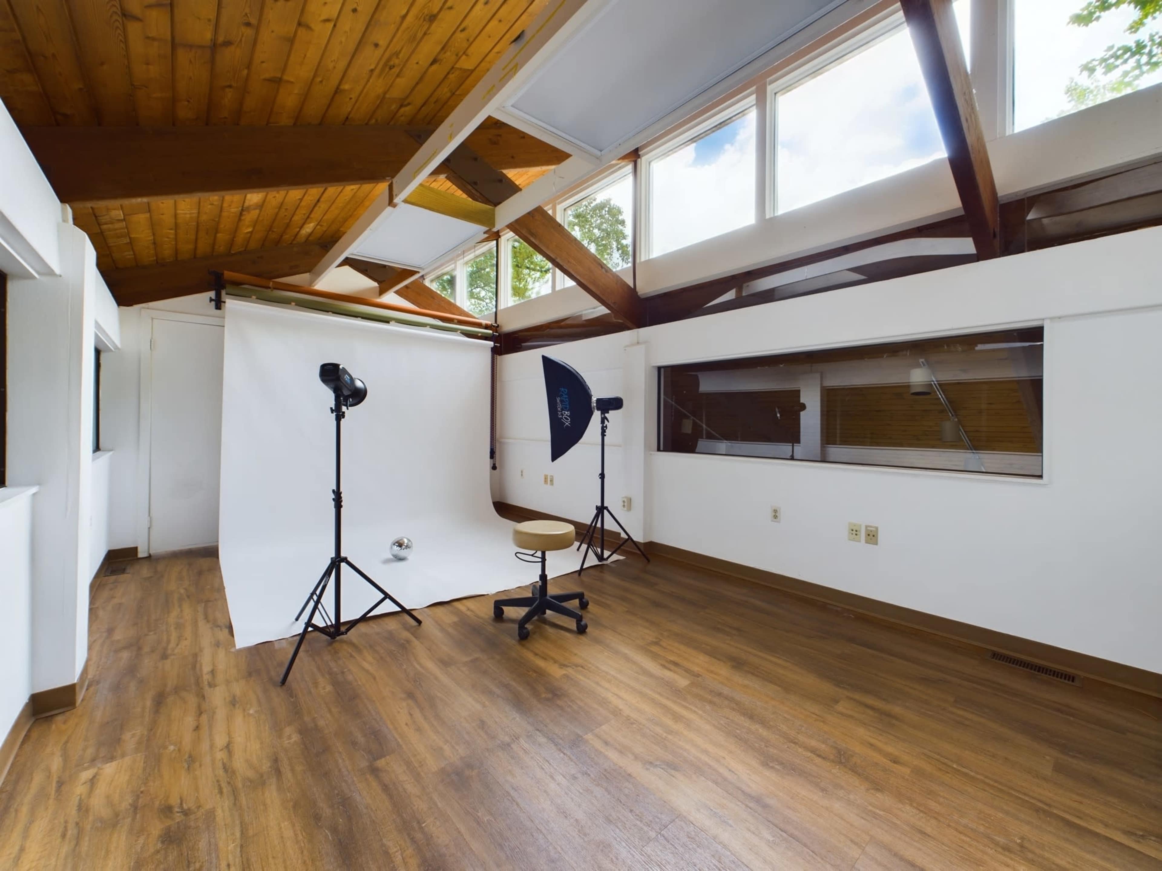 A photography studio with a white backdrop, two lighting setups, a stool, and large windows allowing natural light in.