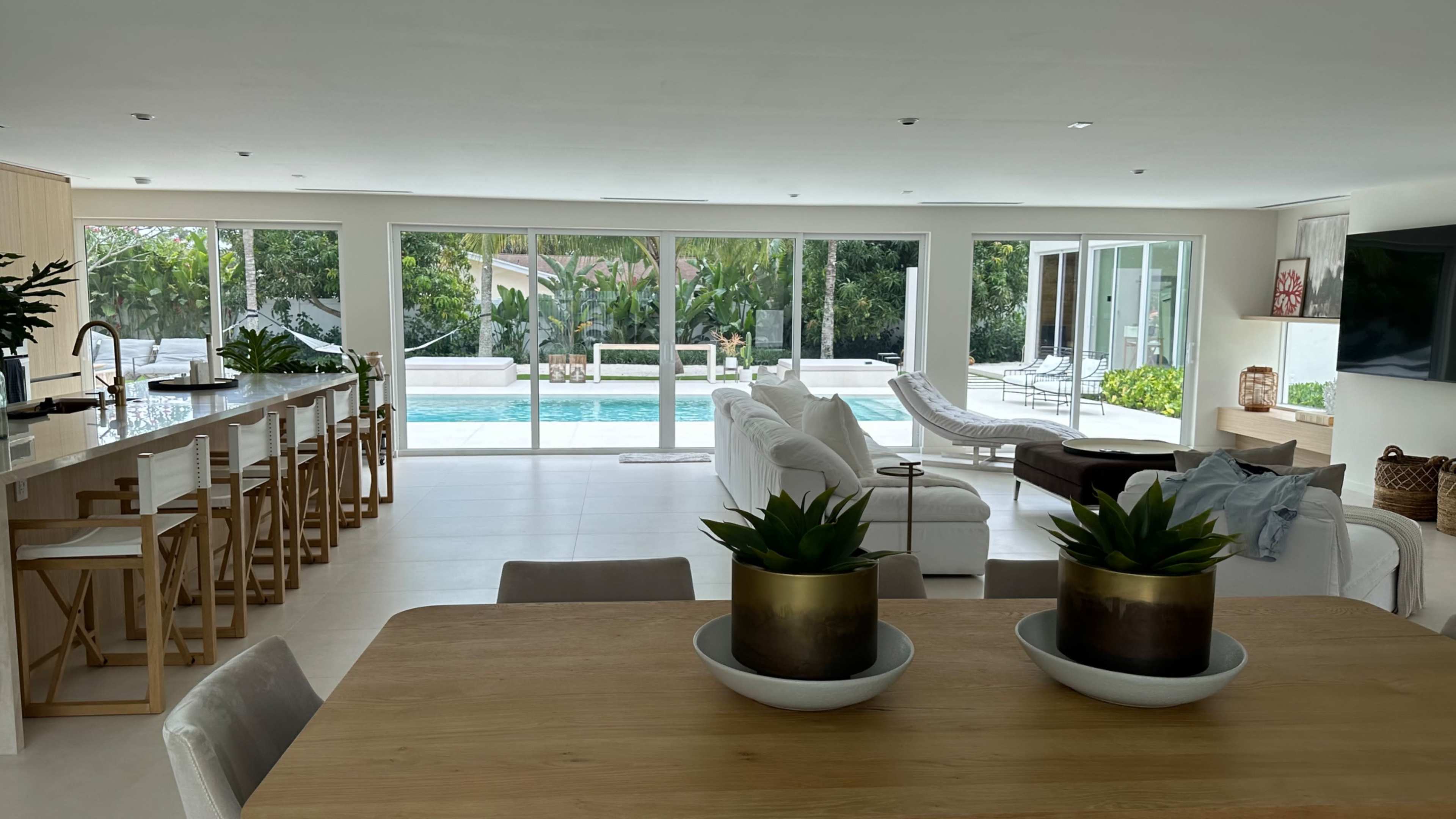 The image shows a modern interior space with a dining area, a seating area, and large glass doors leading to a poolside patio.