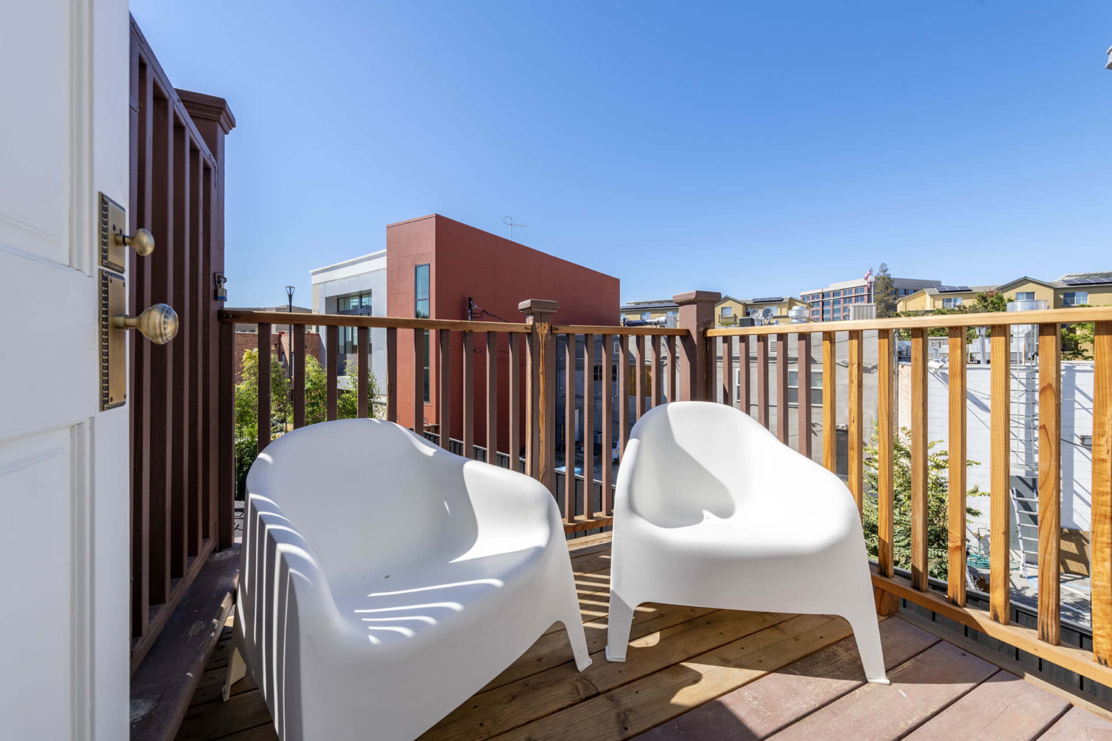 A small wooden balcony enclosed by a railing, featuring two white chairs and a clear blue sky overhead.