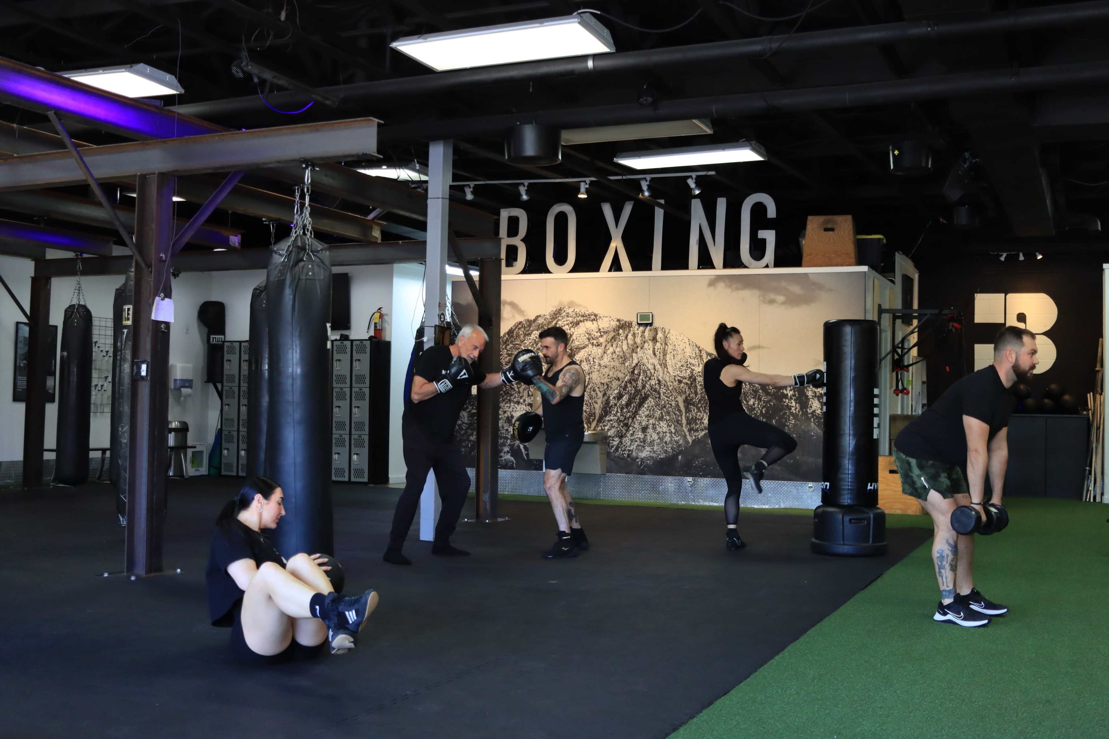 A fitness studio features several individuals engaged in various exercises, including weightlifting, sit-ups, and boxing, with punching bags in the background.