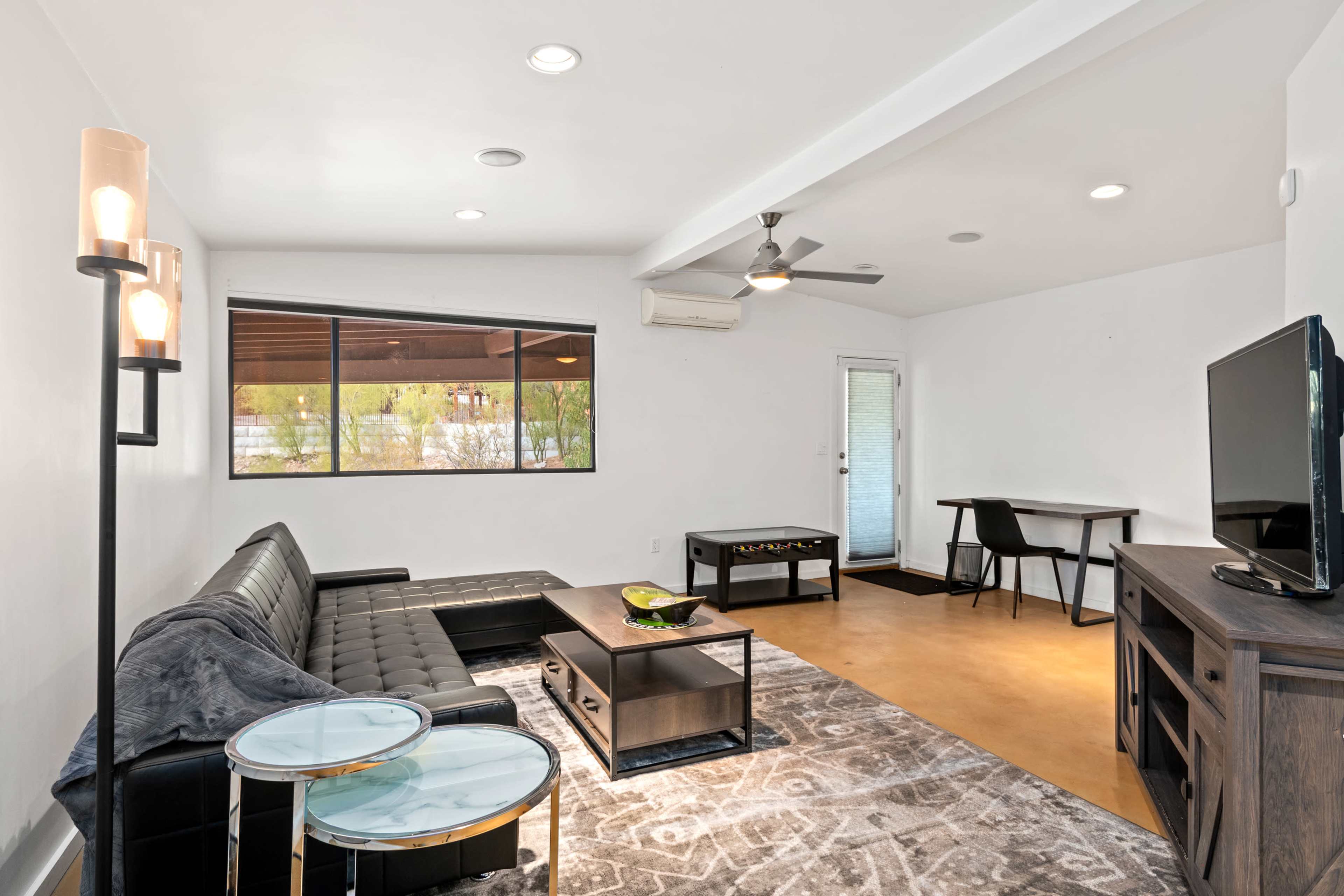 A modern, minimalist living room with a black sofa, a glass side table, a TV cabinet, and a small dining area near a sliding door leading outside.