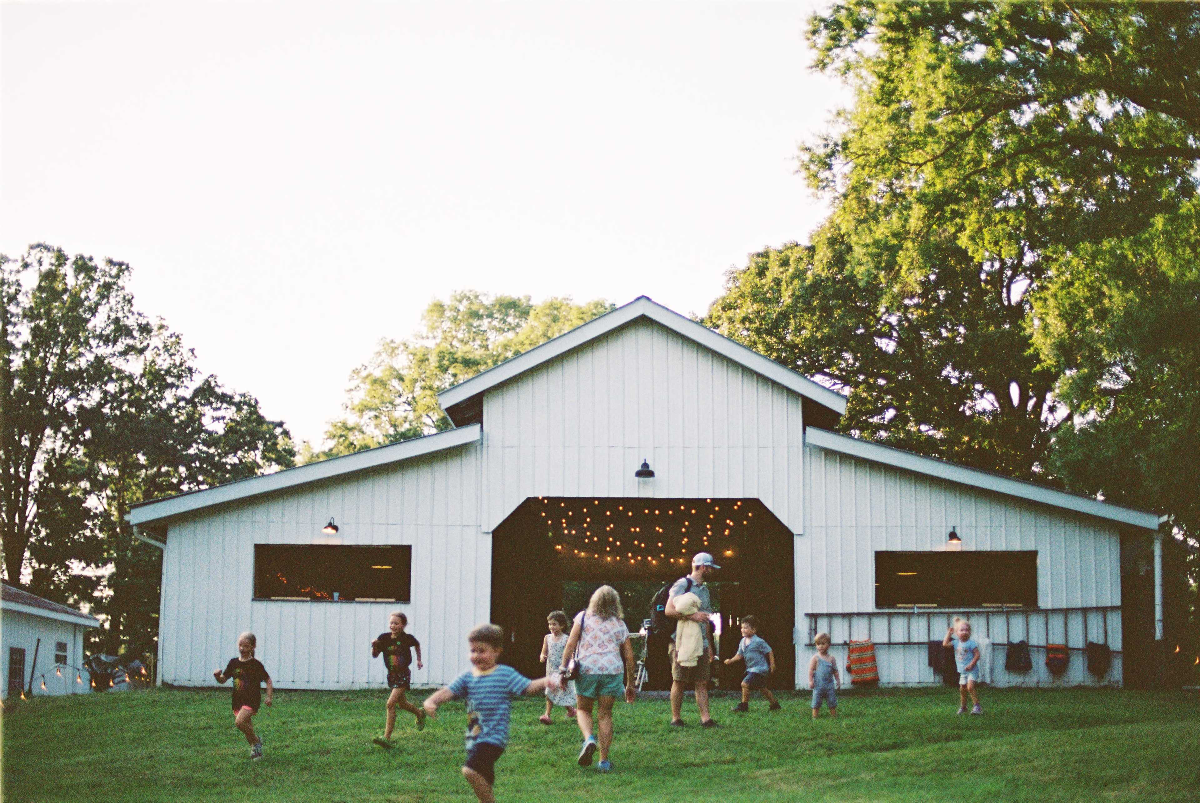 Children run and play in front of a large white barn with open doors, surrounded by trees and string lights.
