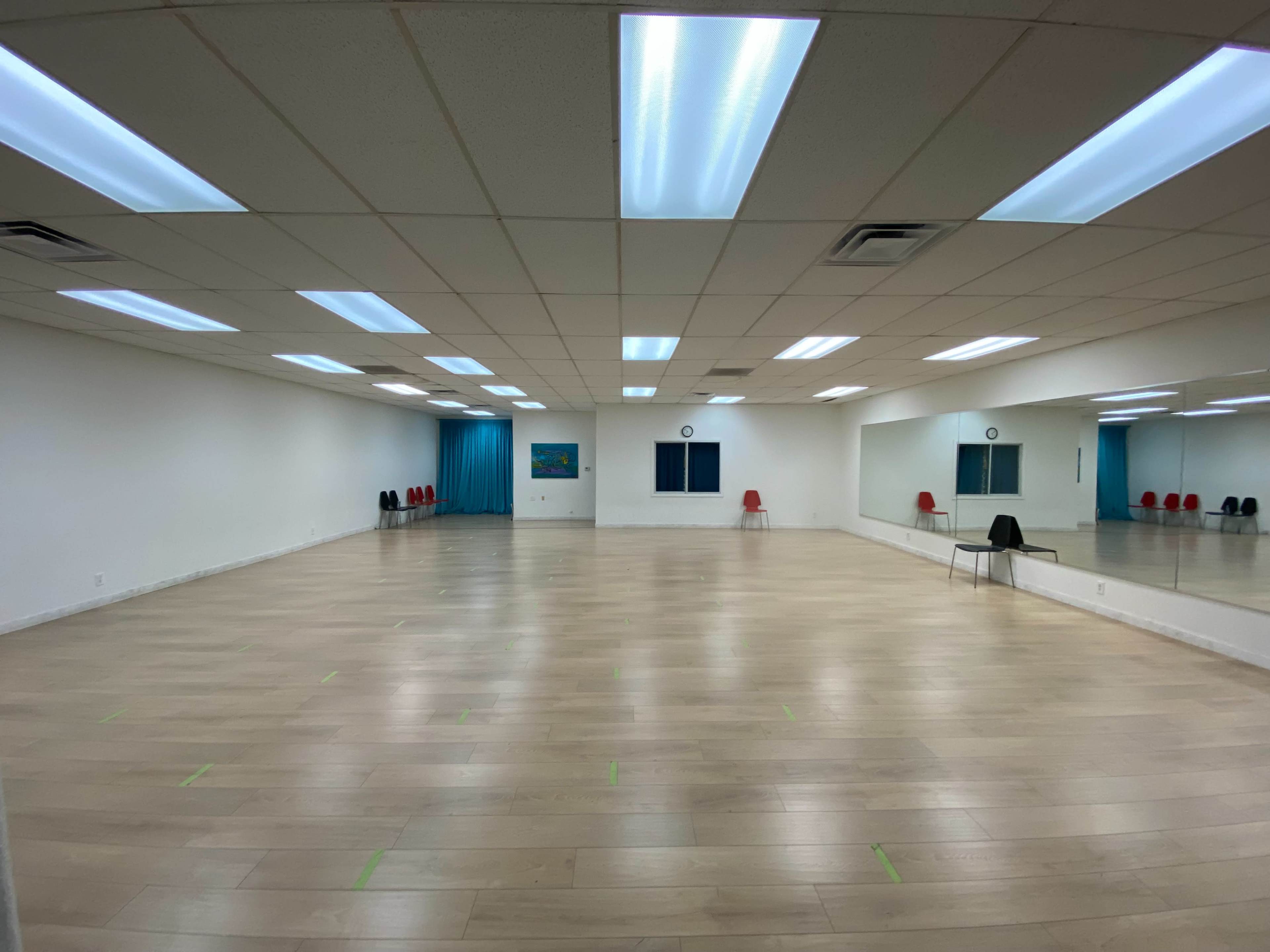 A spacious, well-lit studio with a wooden floor, large mirrors on one wall, and several chairs lined against the opposite wall.
