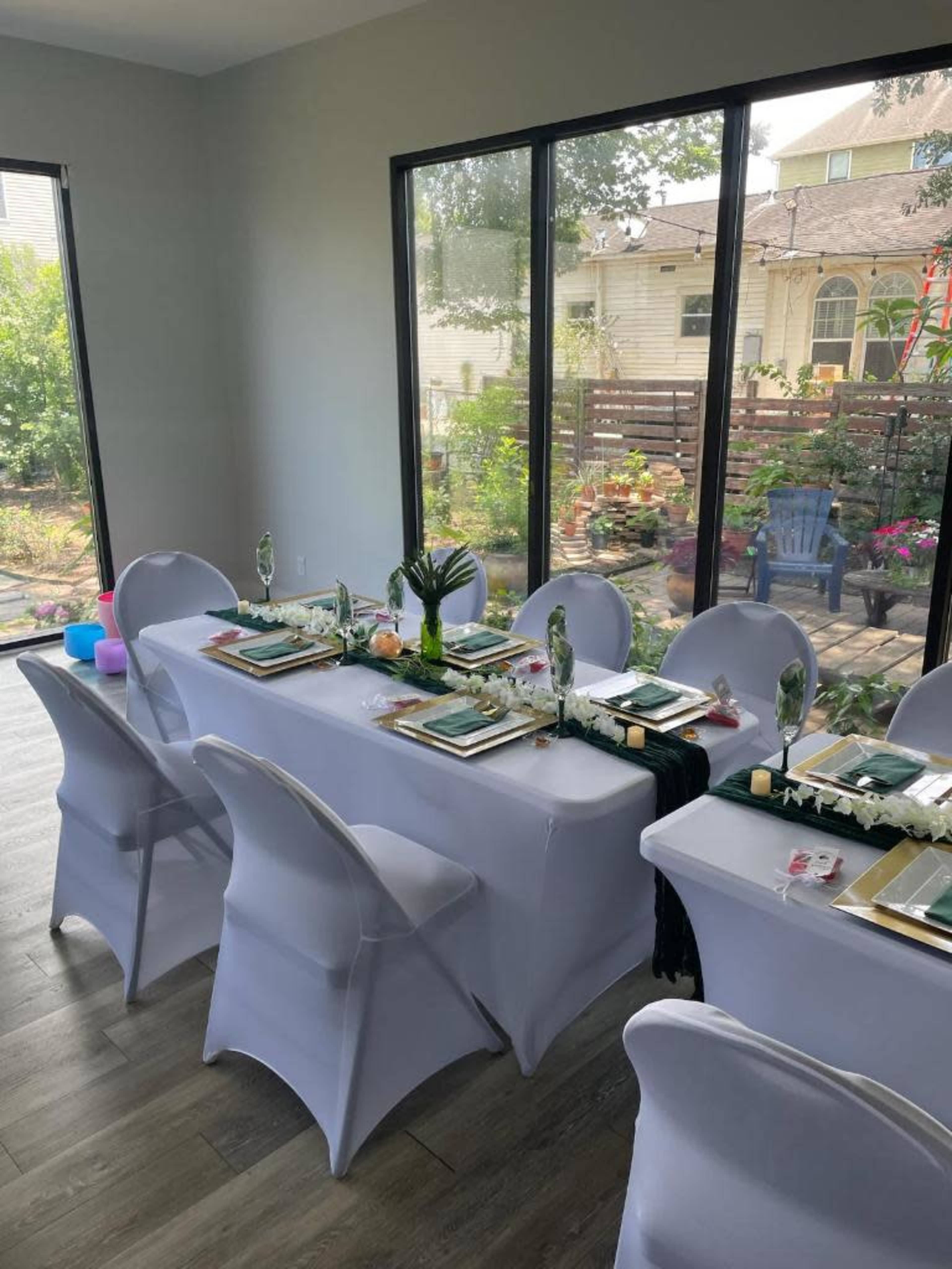 A long dining table is set with plates and utensils, surrounded by white chairs, and facing large windows that overlook a garden.
