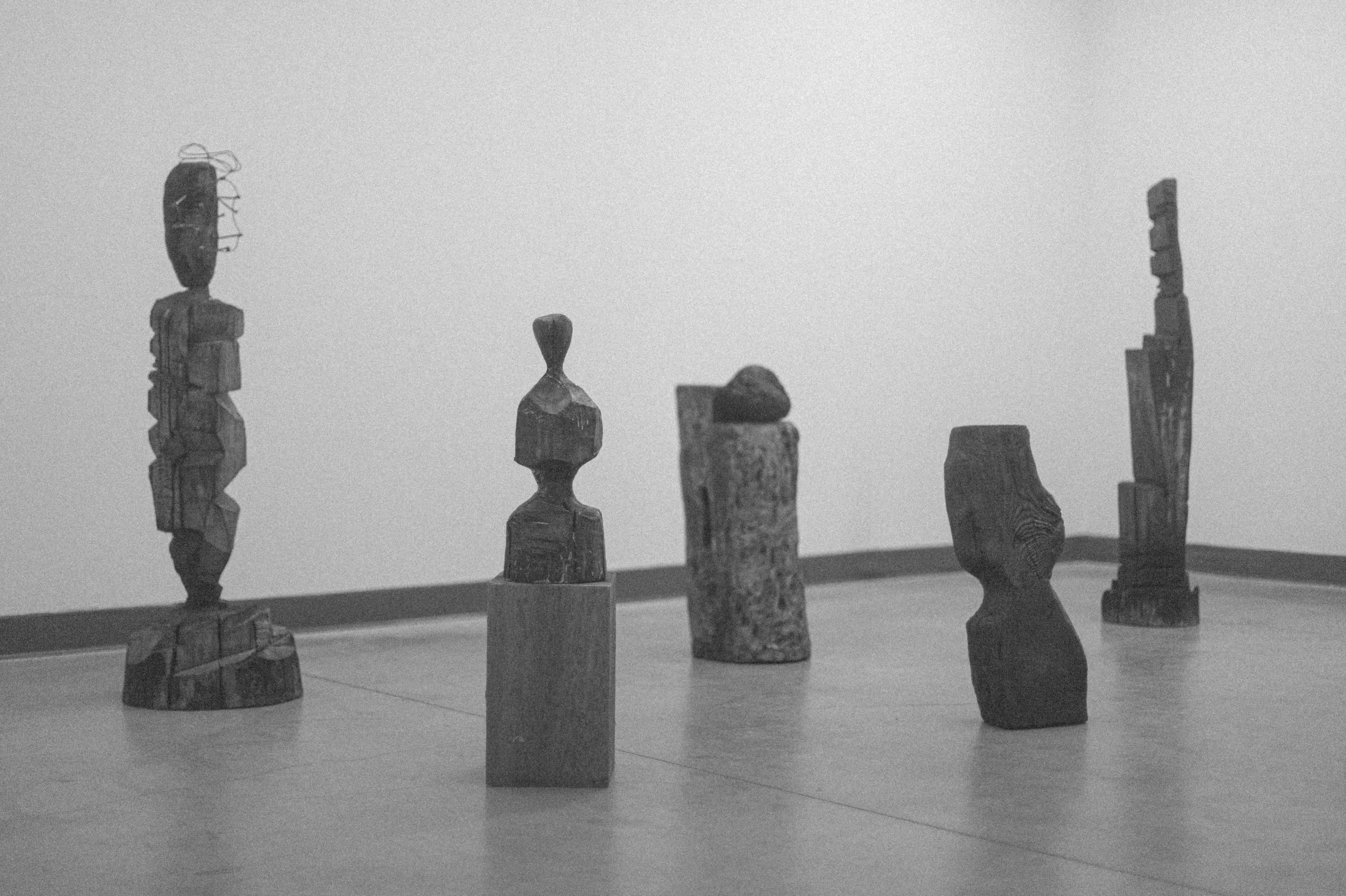 The image features a collection of six abstract wooden sculptures arranged in a minimalist gallery space.