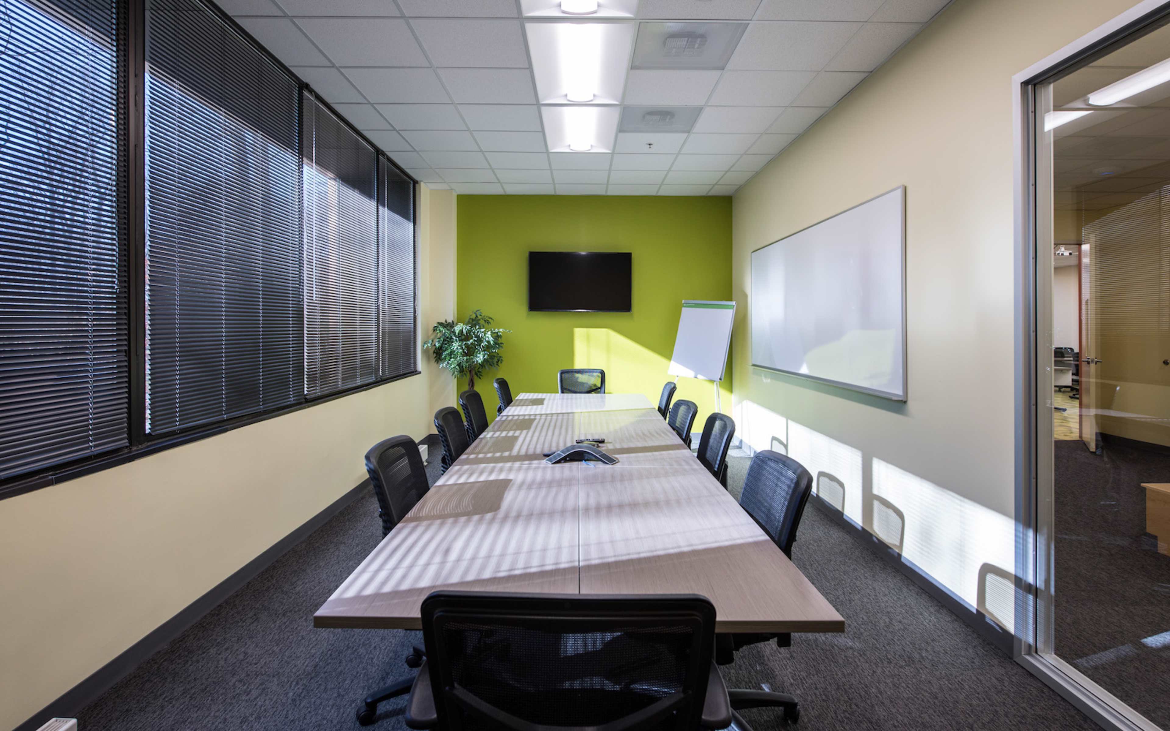 The image shows a meeting room featuring a long table with black chairs, a television on the wall, a whiteboard, and bright green and yellow walls.