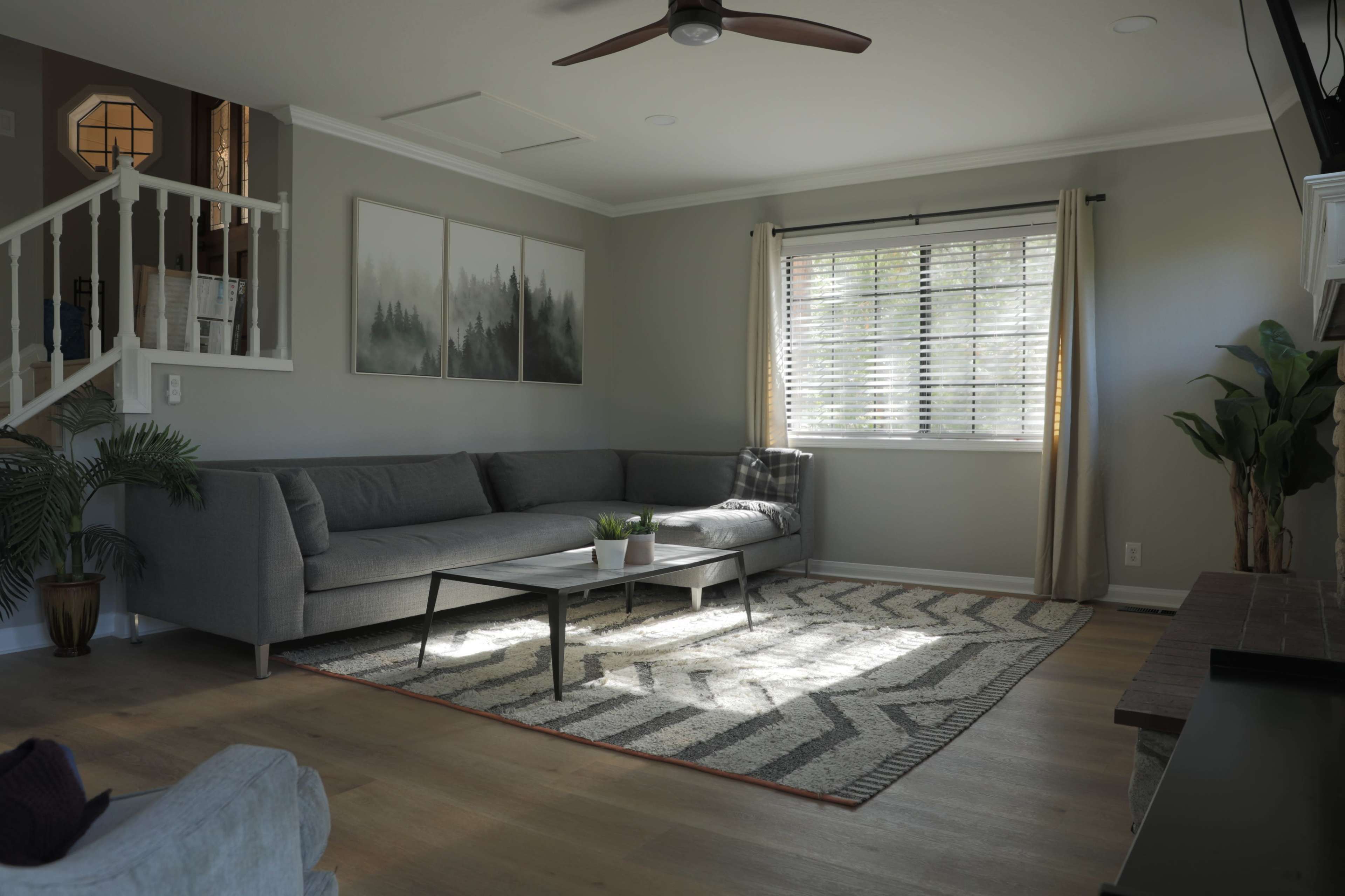The living room features a gray sectional sofa, a coffee table, and a patterned rug, with large windows allowing natural light to illuminate the space.