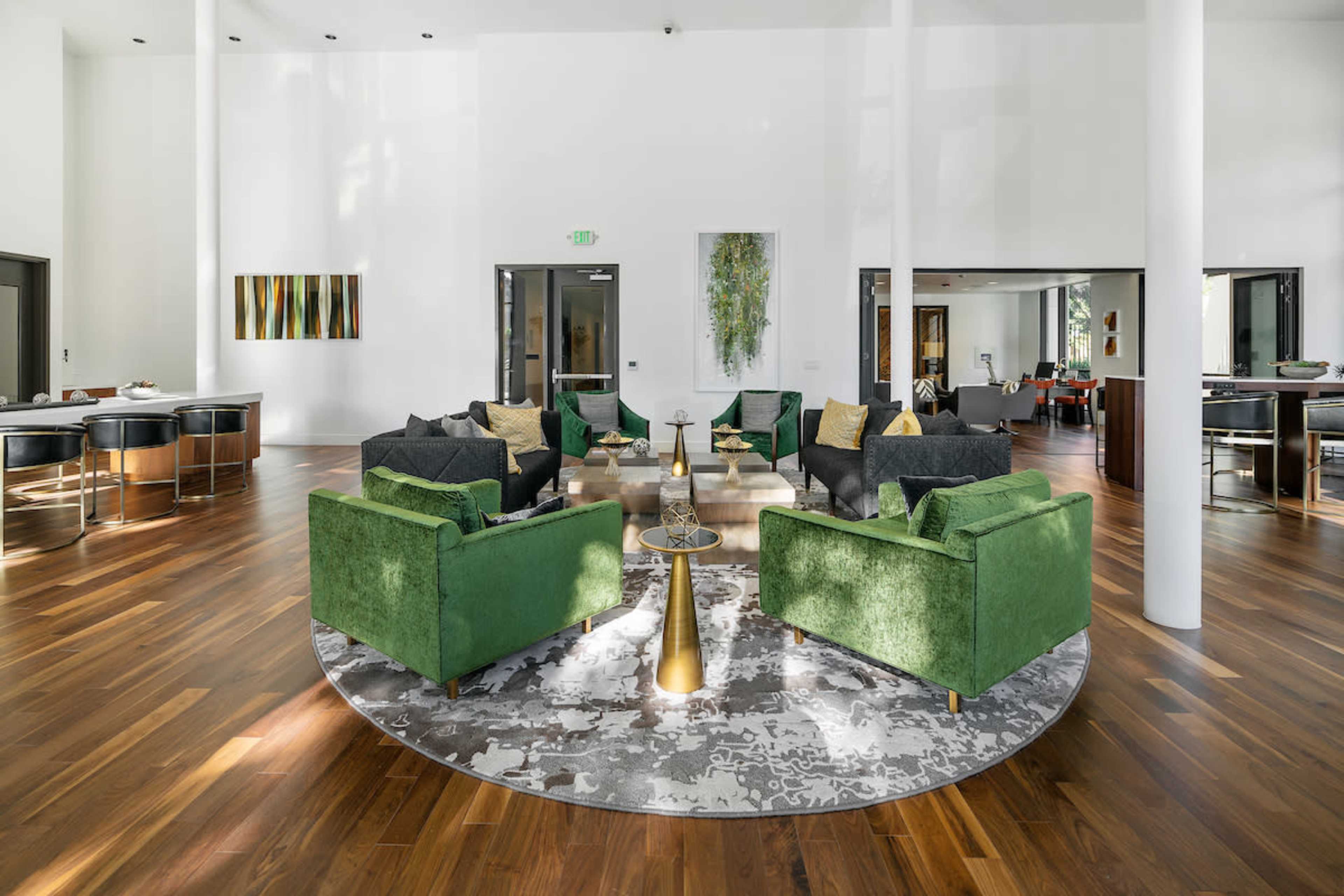 A spacious room features circular arrangements of green armchairs on a patterned rug, surrounded by large windows and wooden flooring.