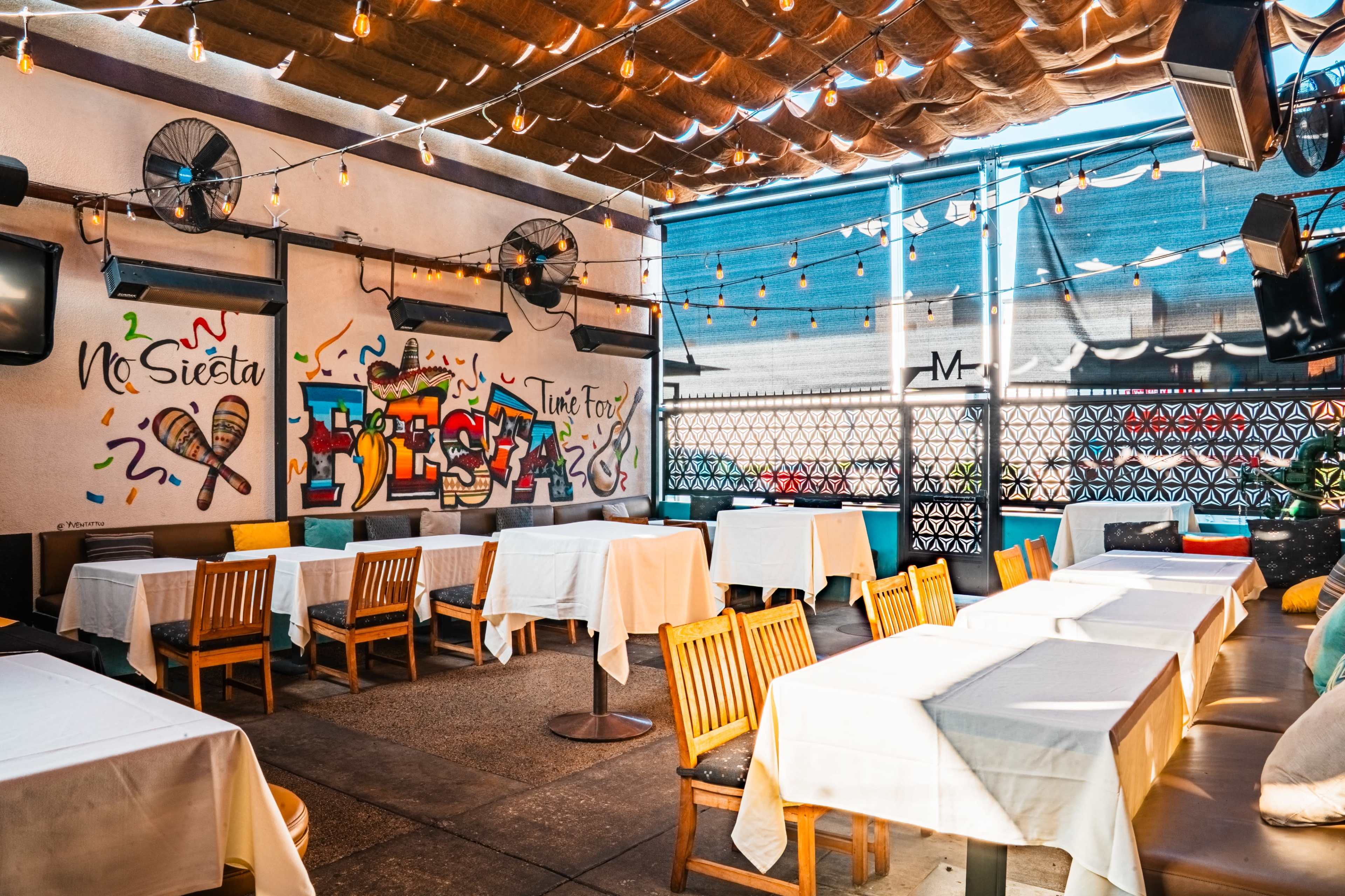 The image shows an empty restaurant patio with wooden tables covered in white tablecloths and a colorful mural on the wall.