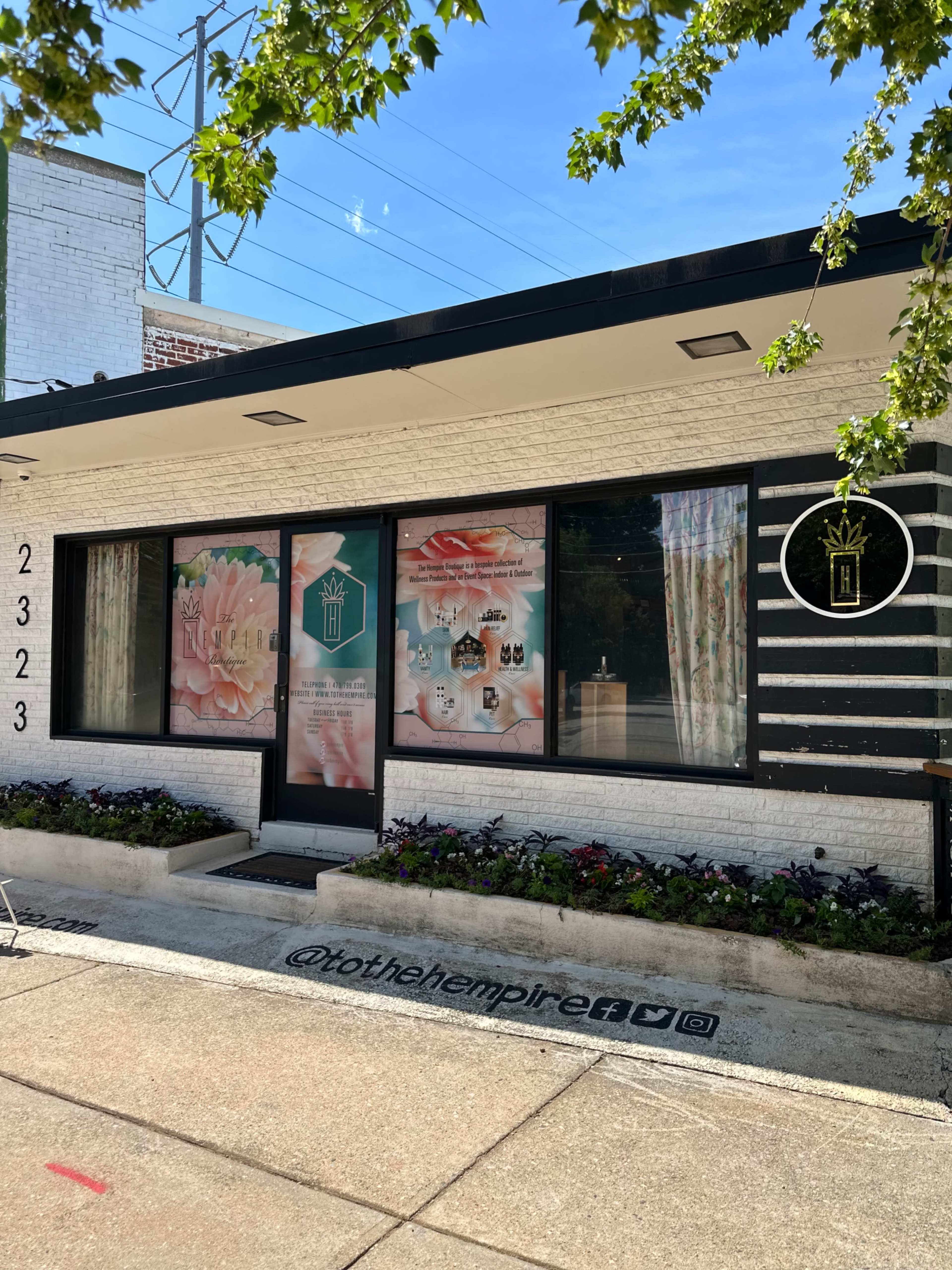 The image shows a storefront with large windows displaying floral graphics and products, situated on a city street with planters along the sidewalk.