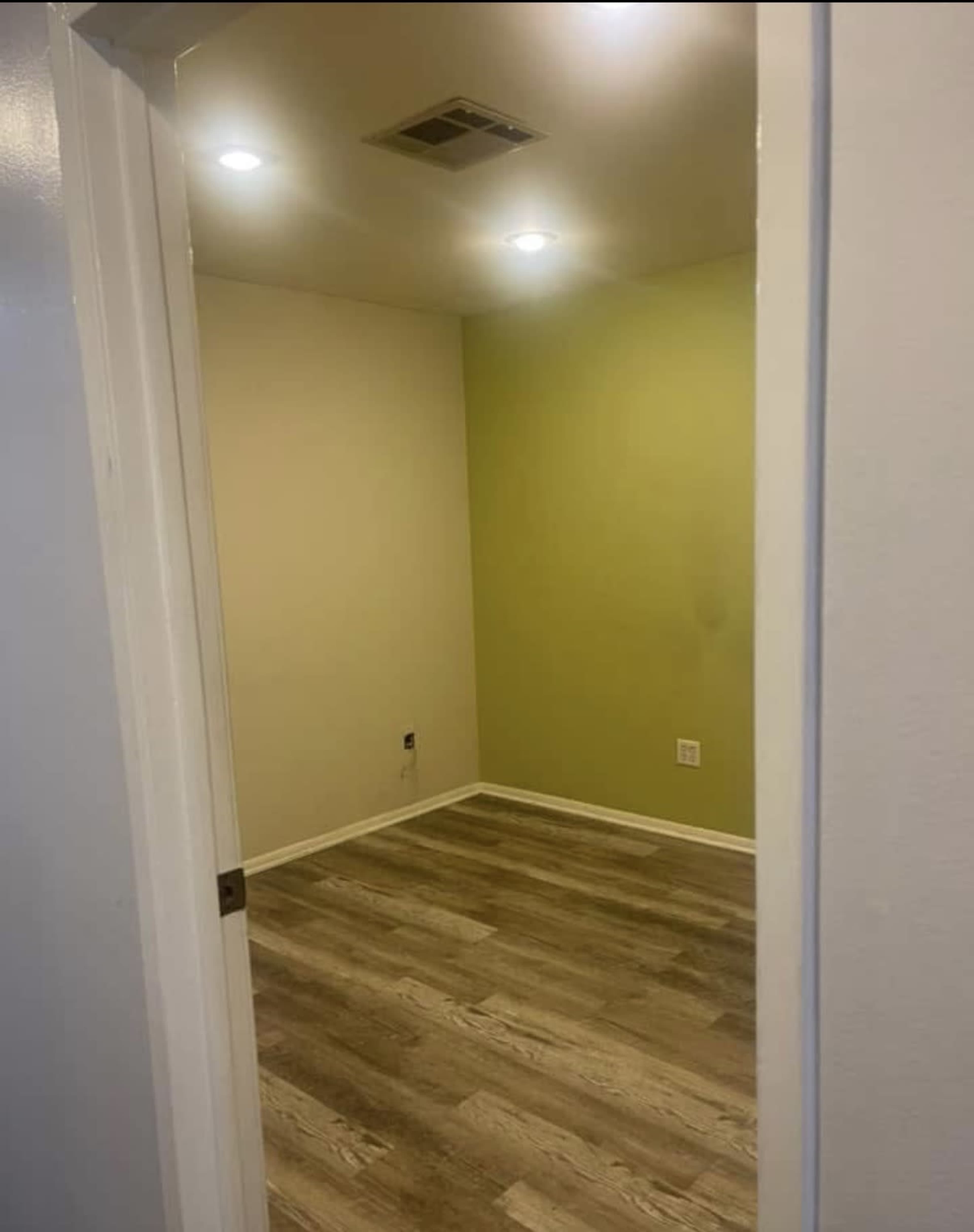 The image shows a small, empty room with a green accent wall and hardwood flooring, viewed from an open doorway.