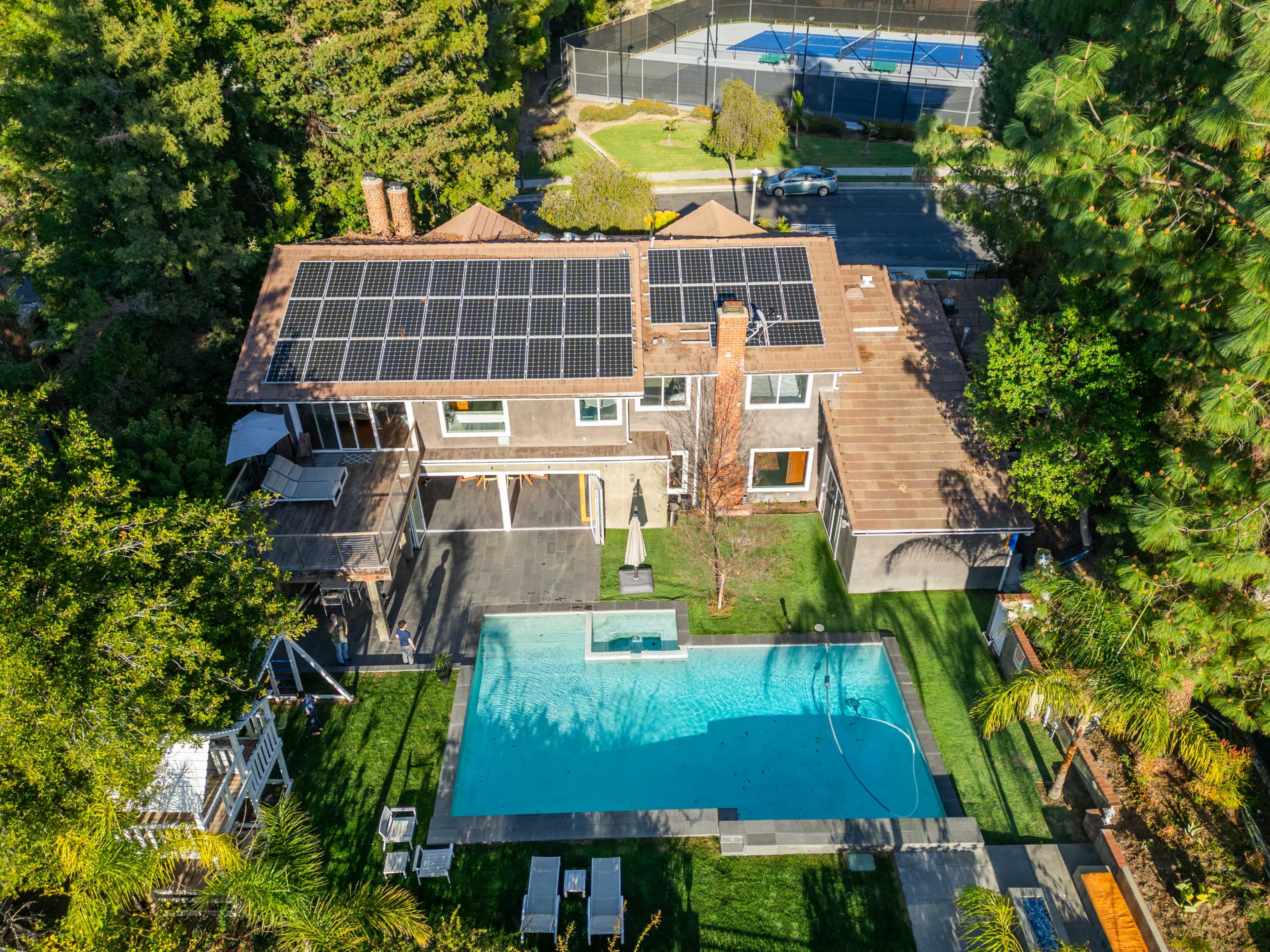 A modern two-story house with a swimming pool and solar panels on the roof is surrounded by trees and a tennis court nearby.