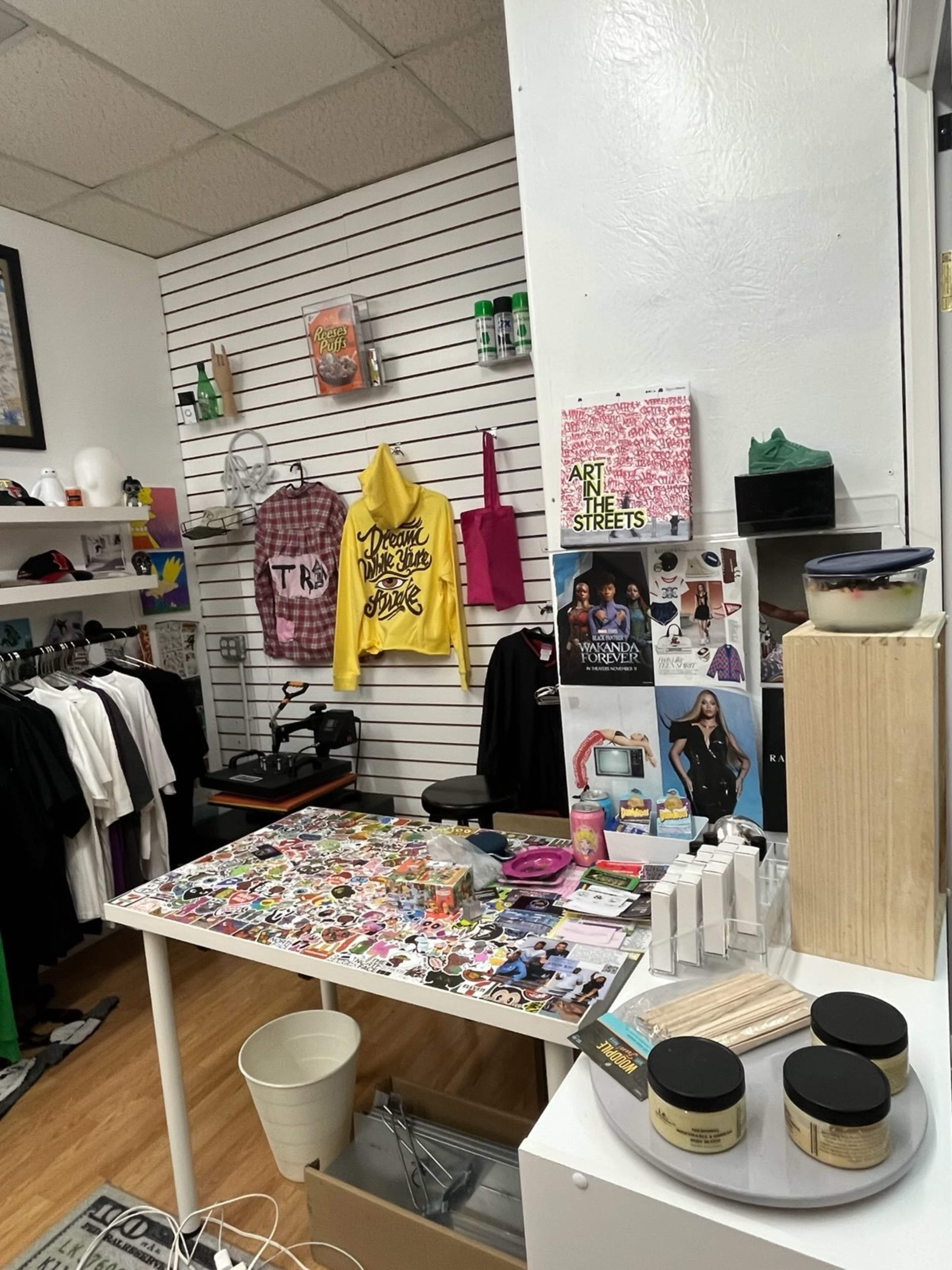 The image shows a small retail space with shelves displaying clothing, accessories, and various art items, along with a cluttered table covered in stickers and other merchandise.