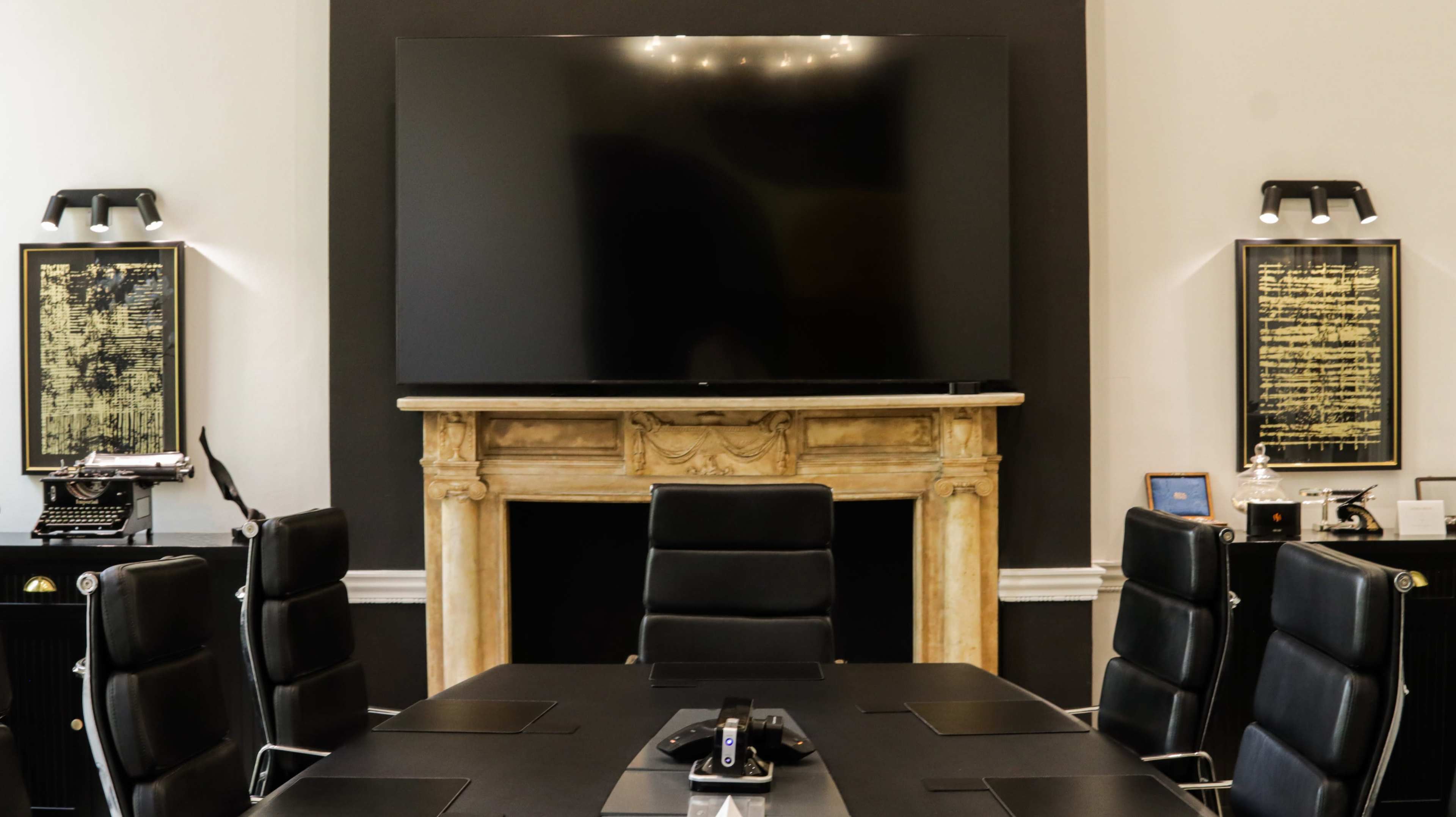 A modern conference room features a large flat-screen TV mounted above a decorative fireplace, with black leather chairs arranged around a rectangular table.