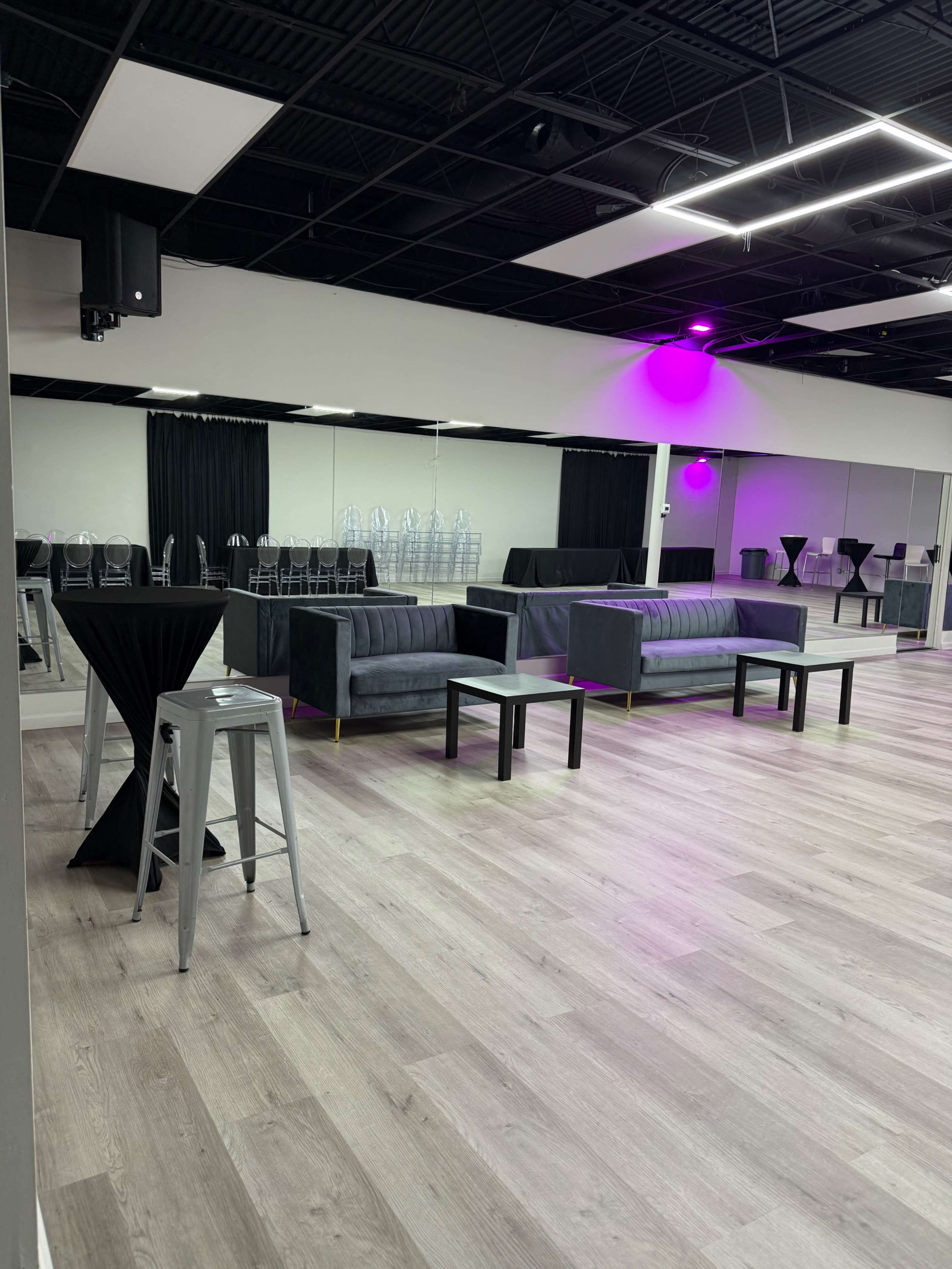 The image shows a spacious room with a light-colored wooden floor, featuring black couches, tables, and clear acrylic chairs arranged in front of a large mirror.