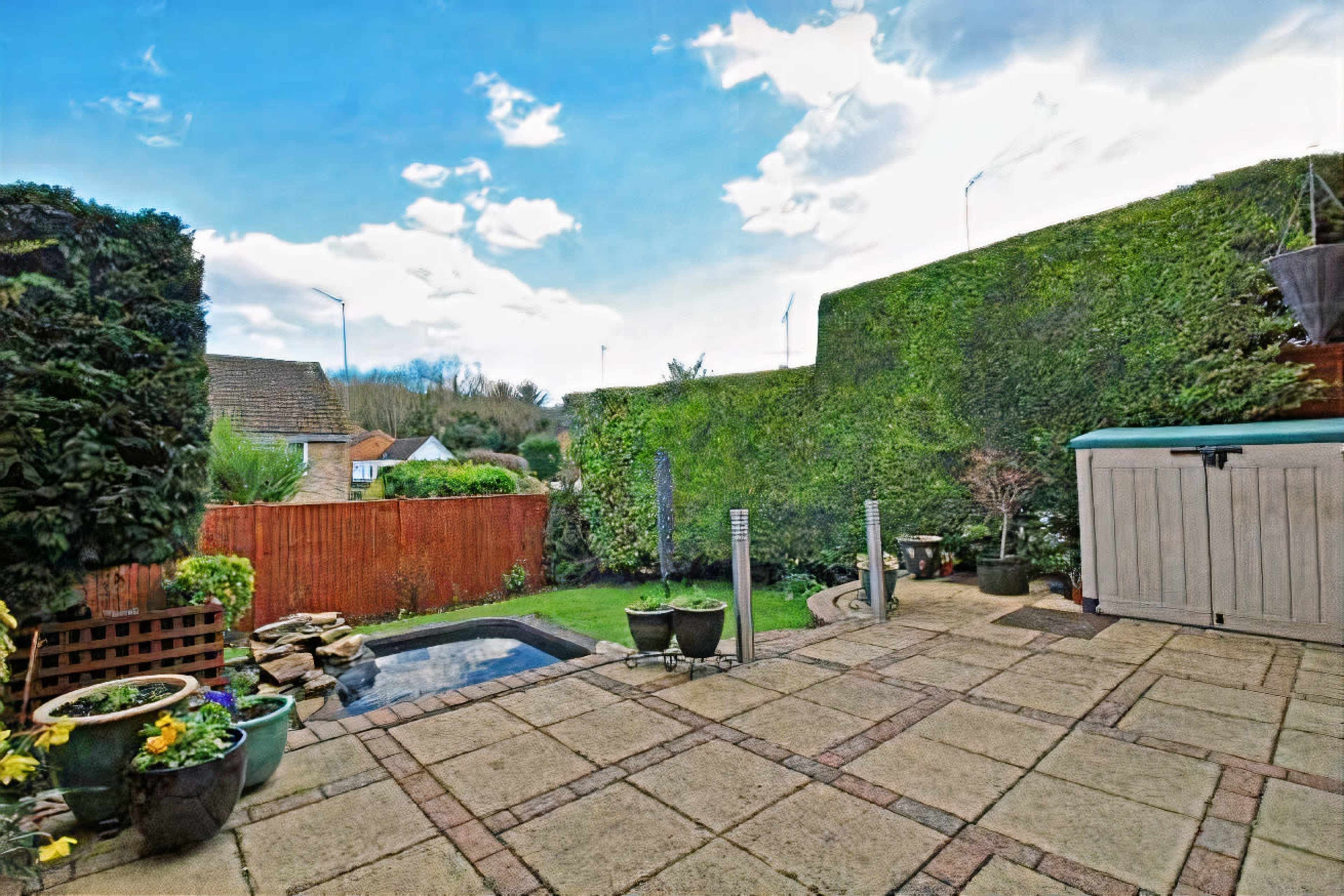 A paved outdoor patio features a small pond, planters, and a garden shed, surrounded by trimmed hedges and a wooden fence.