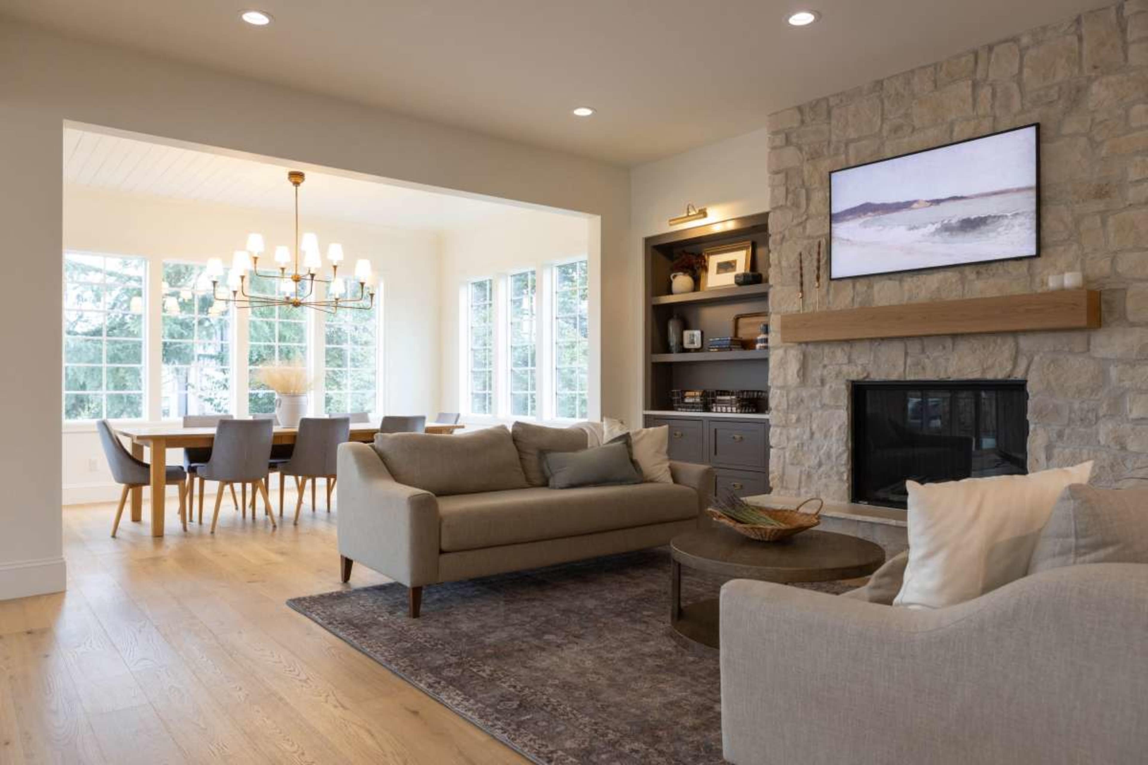A cozy living room features a stone fireplace, modern furniture, and a view into a dining area with a large table and chandelier.
