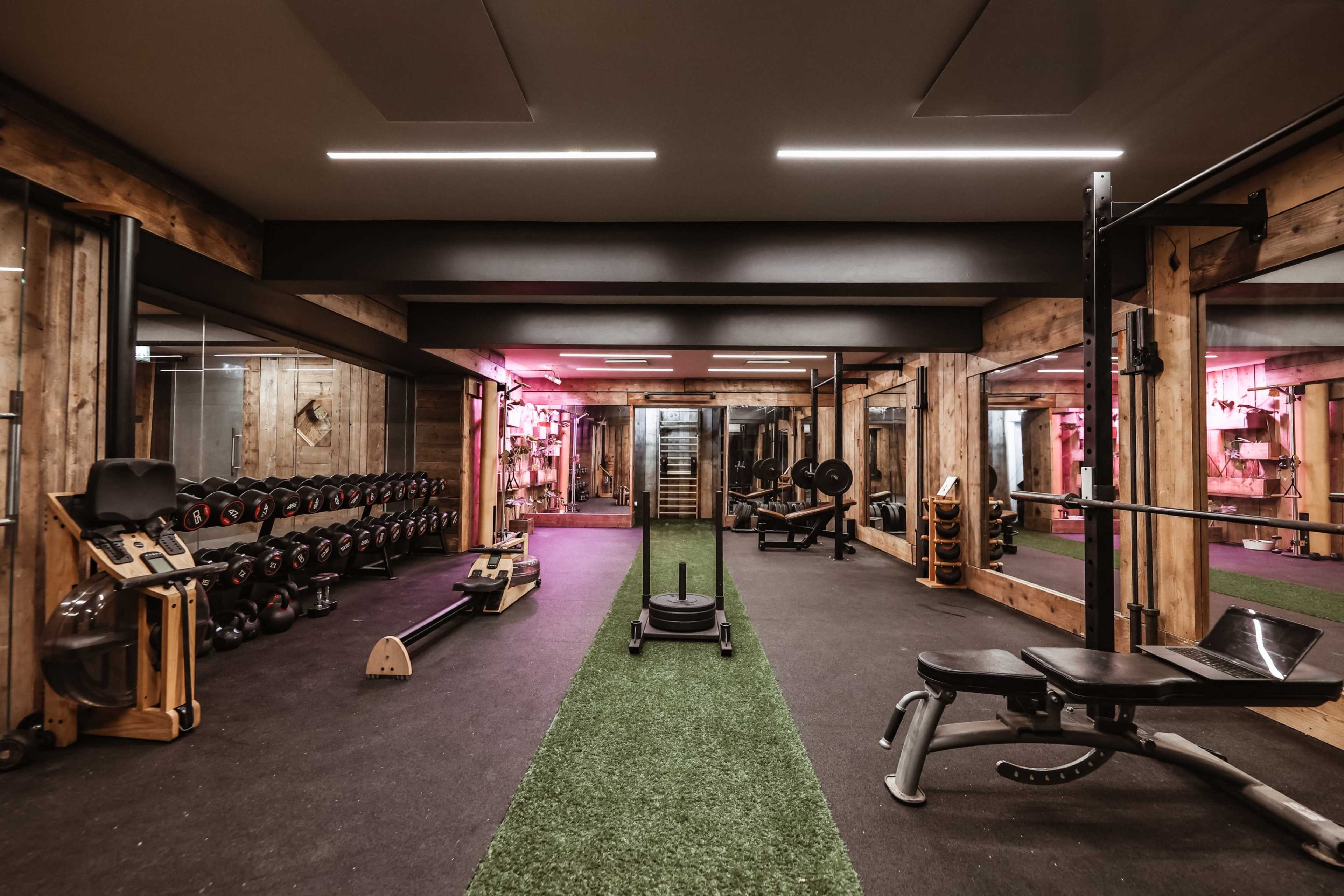 The image depicts a well-equipped gym featuring various weightlifting equipment, a sled track, and a turf area, with wooden walls and modern lighting.