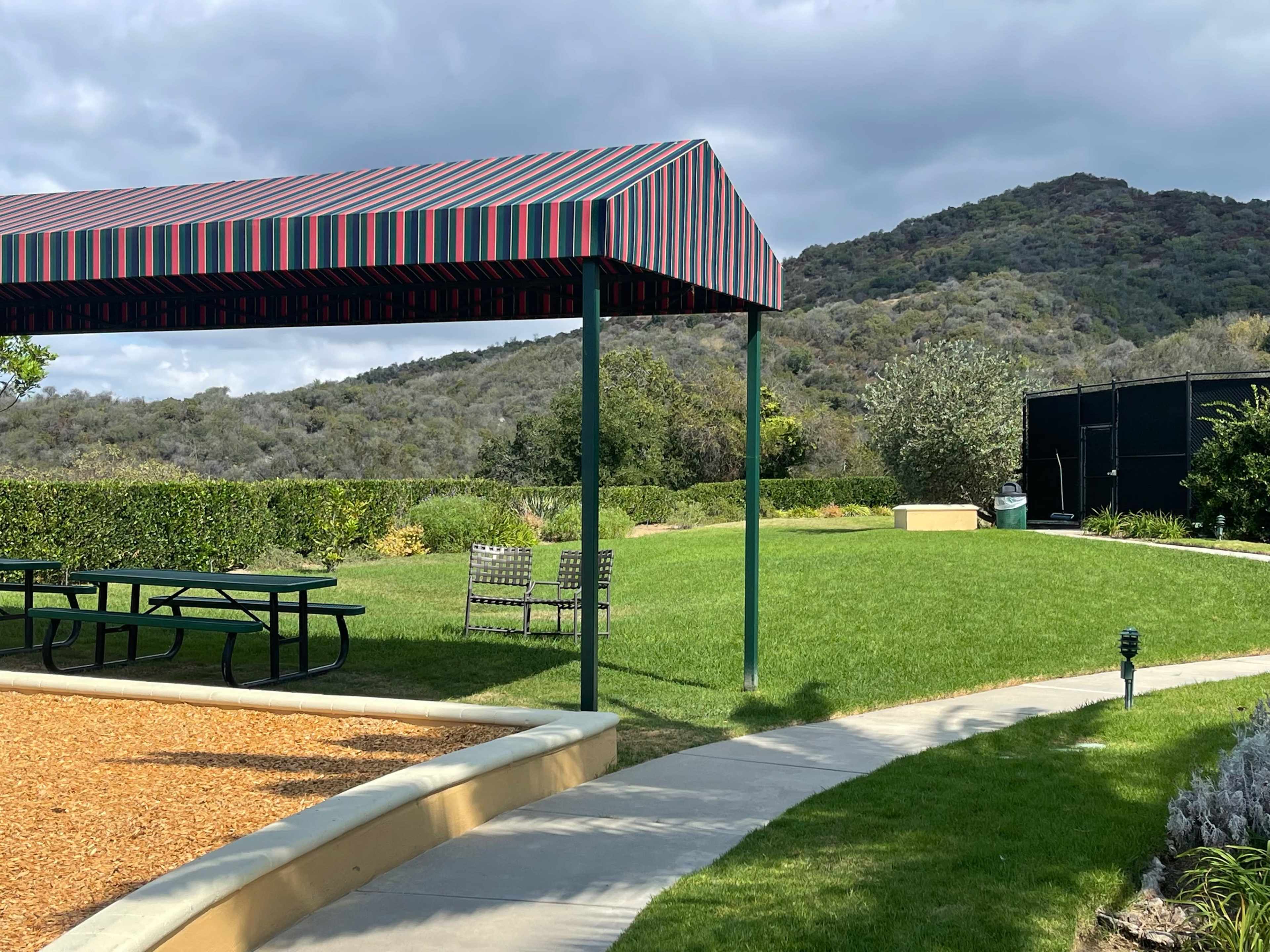 A shaded picnic area with a striped canopy is set beside a green lawn and a path leading to a garden and hillside in the background.