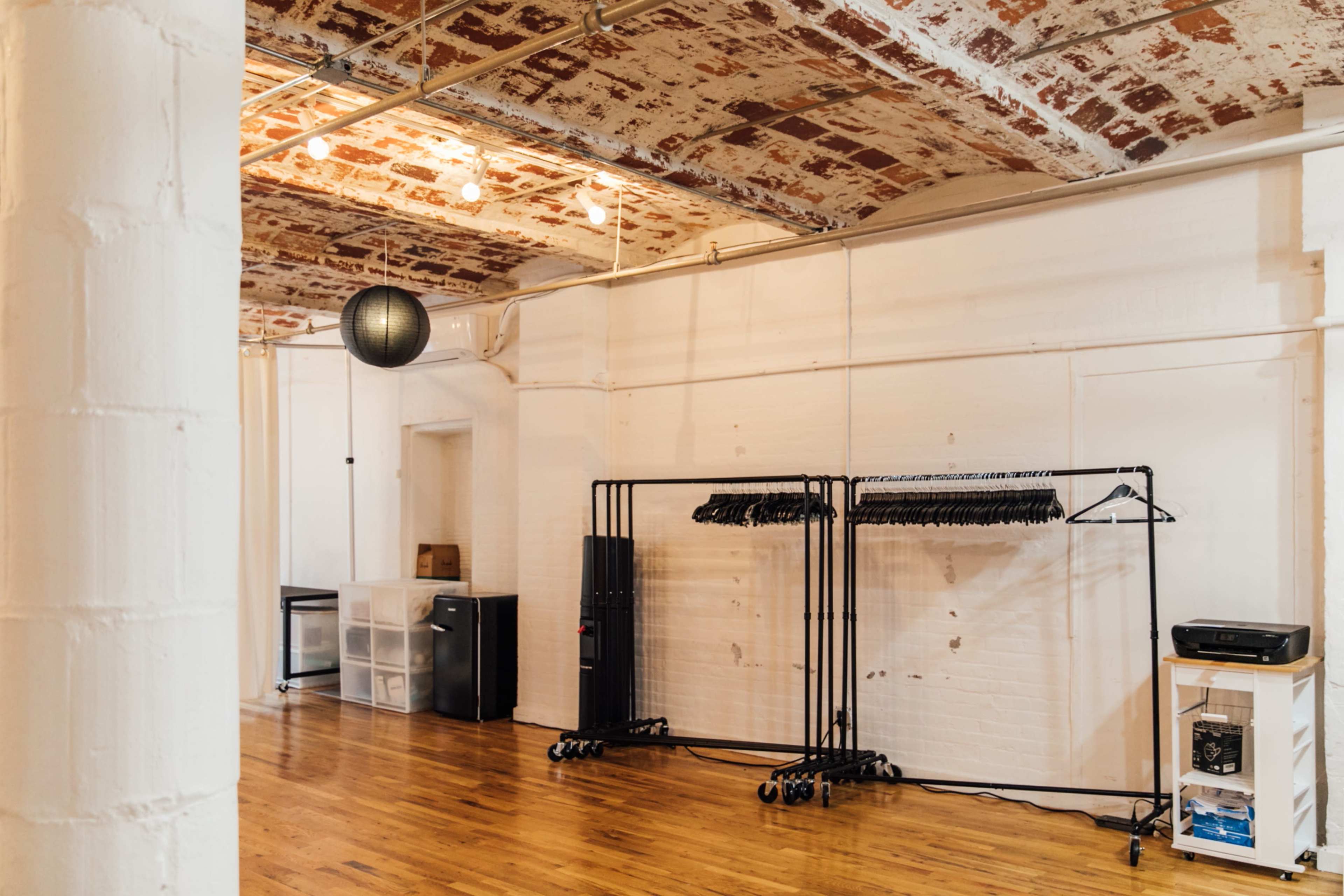 An empty room featuring exposed brick walls, wooden flooring, and a black hanging light fixture, along with clothing racks and storage units.