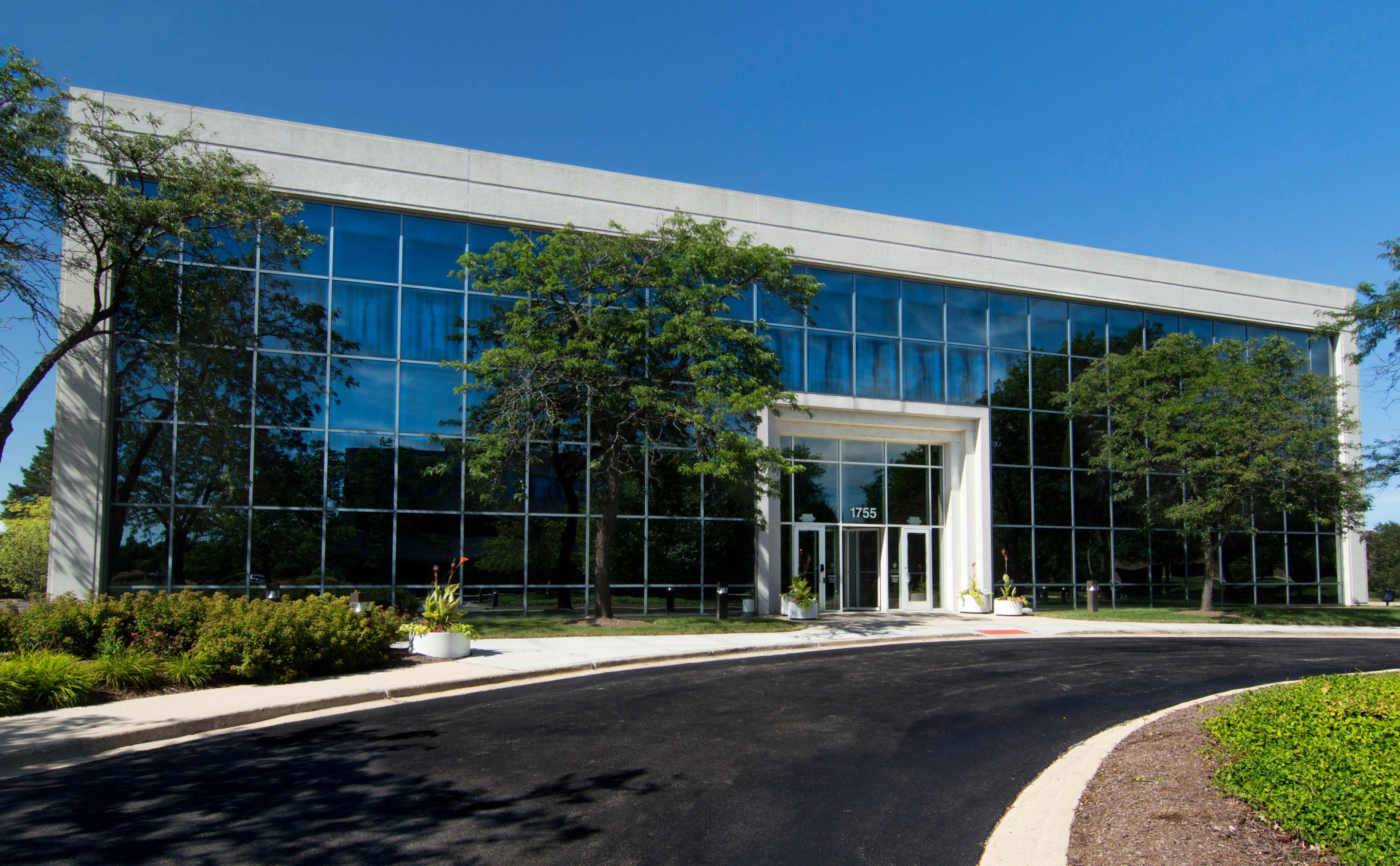 A modern office building with large glass windows is set against a clear blue sky, surrounded by greenery.